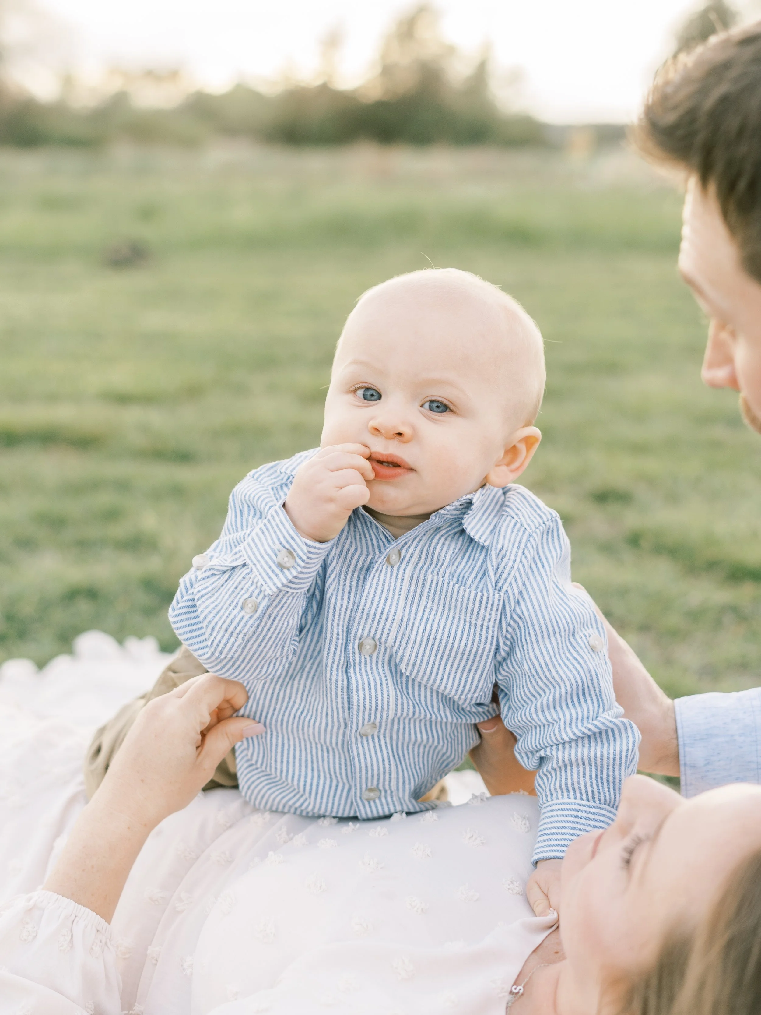 A baby boy with blue eyes sitting on an adult's lap outdoors, touching his lips with one hand, while an adult gently holds him.