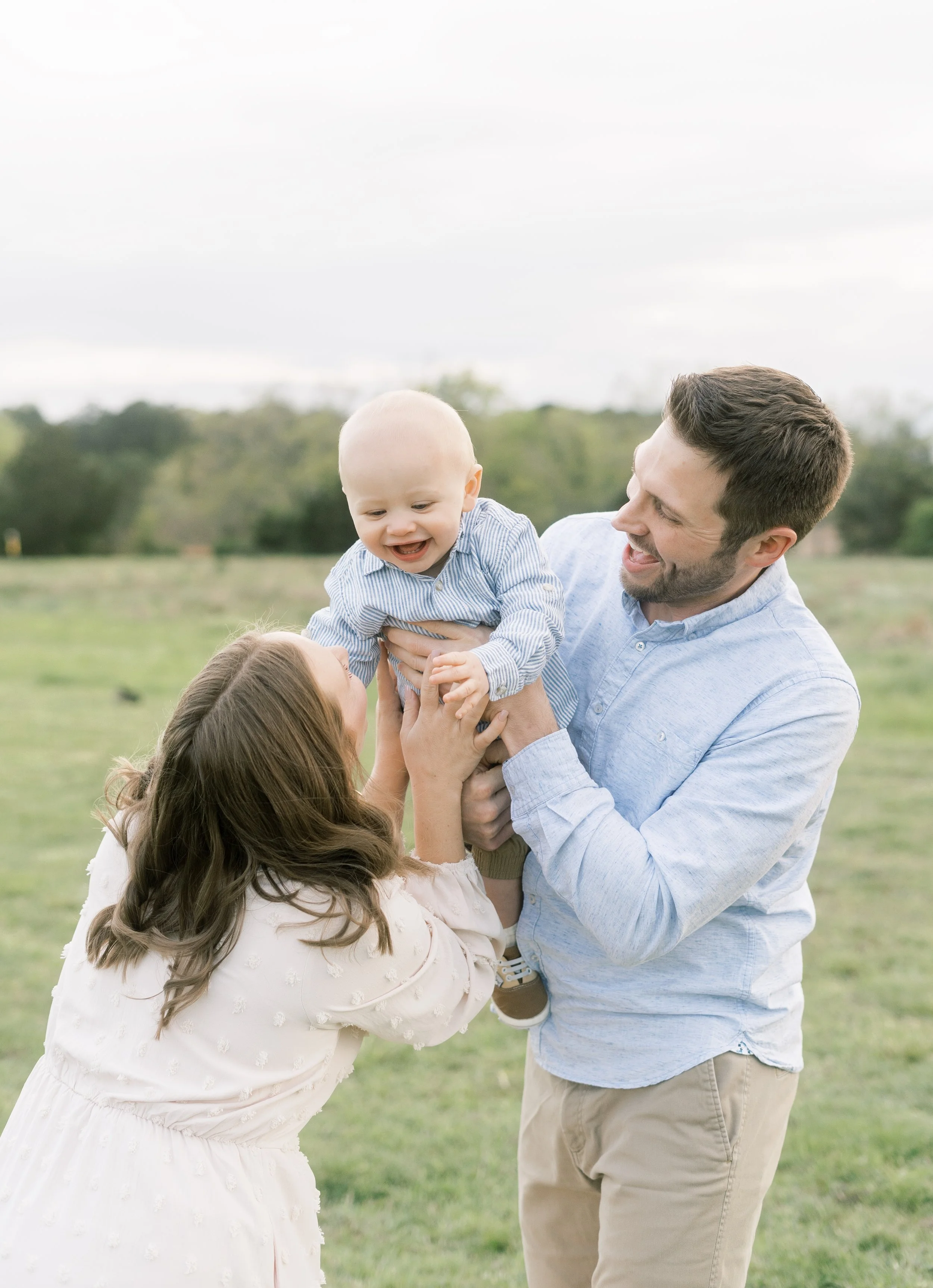 A family of three playing outside in a grassy field, with the father lifting the baby boy in the air while the mother reaches up to hold his hand, all smiling and enjoying the moment.