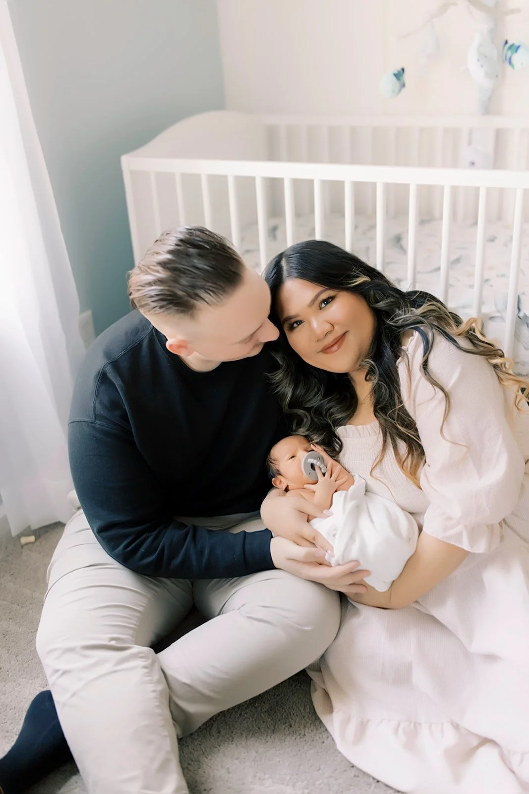 A happy family moment with a woman, man, and newborn baby in a nursery, with the mother holding the baby and both parents looking lovingly at the baby.