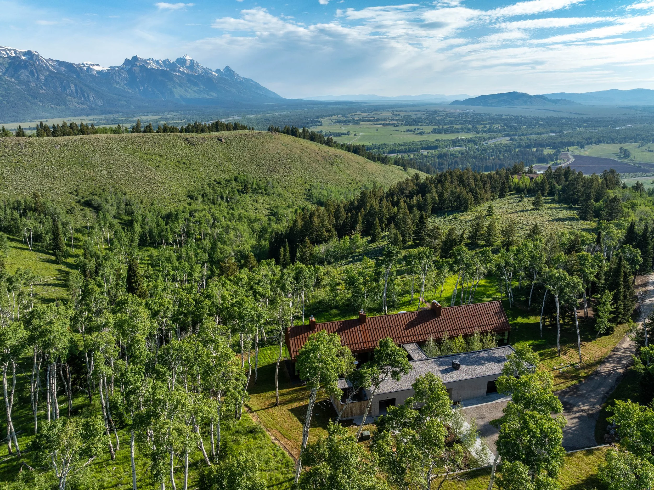 Aerial photo of a house with a red roof and a concrete garage, surrounded by trees and rolling green hills, with mountains in the background under a partly cloudy sky.