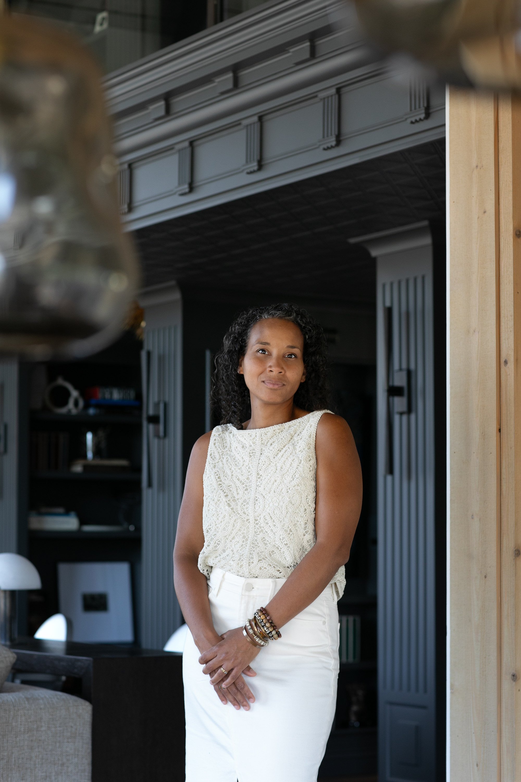 A woman with black, curly hair standing in a modern indoor space, wearing a sleeveless lace top and white pants, with her hands clasped in front of her, looking at the camera with a slight smile.