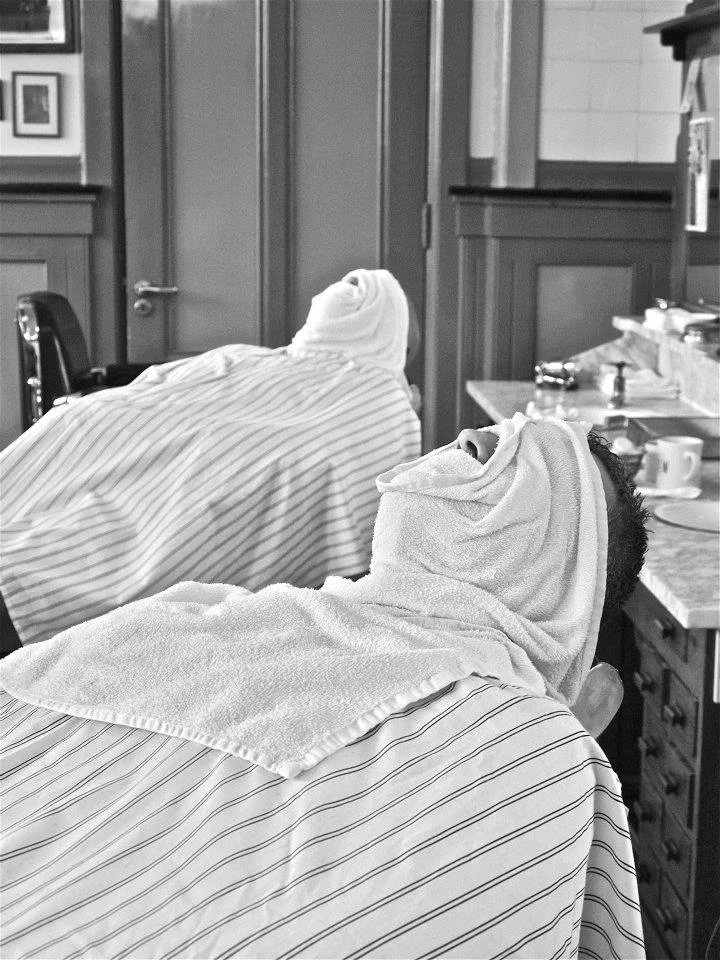 Black and white photo of two men lying back in barber chairs with towels covering their faces, surrounded by traditional barbershop furnishings. Sligo best hot towel shave haircut