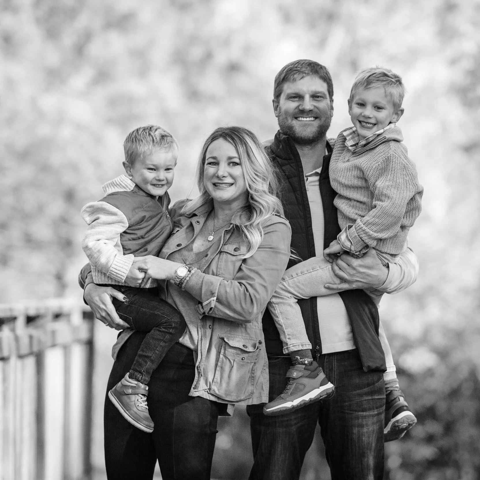 Black and white photo of a smiling family in an outdoor setting. A woman and a man stand close together, each holding a young child. The family appears happy and relaxed.