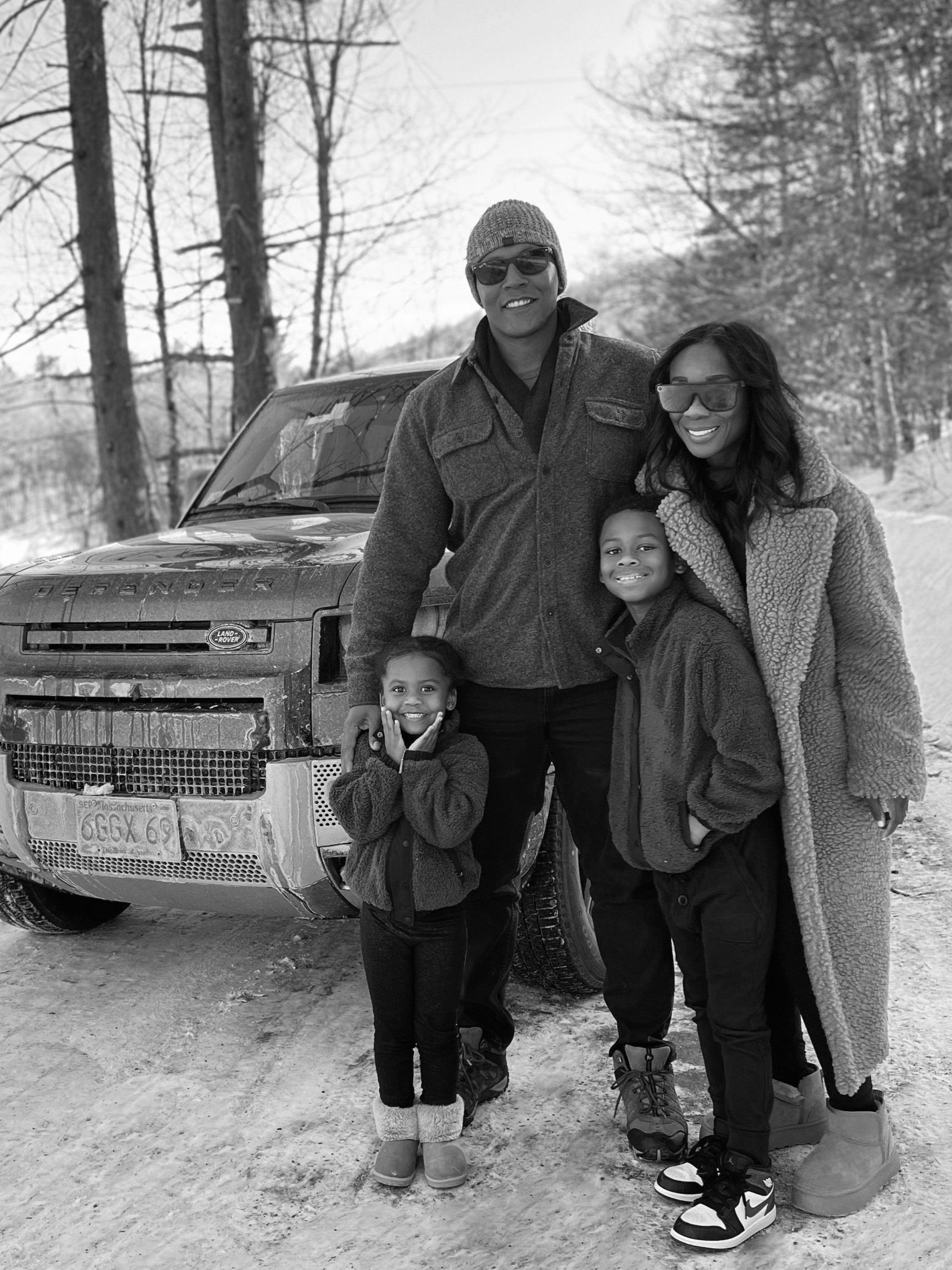 A family of four standing in front of a truck in a snowy forest, all wearing winter clothing.