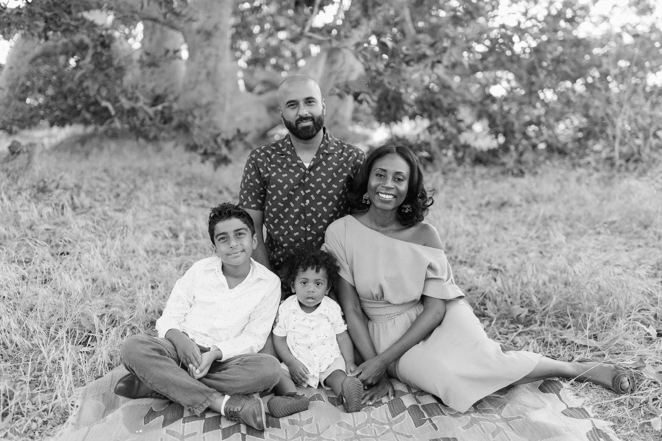 Black and white photo of a family sitting on a blanket outdoors. There are two adults and two children, one of whom is a toddler. They are surrounded by trees and grass.