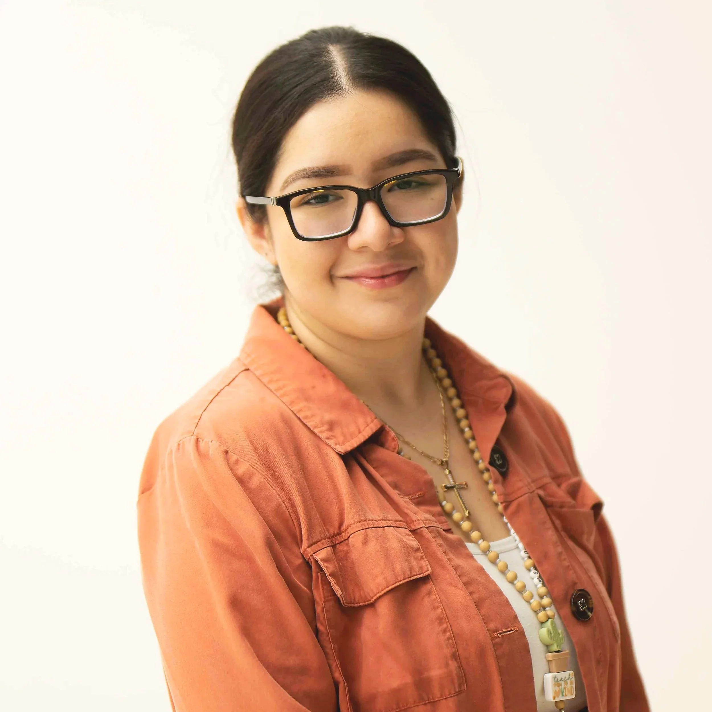 A person wearing black glasses, a white shirt, and a rust-colored cardigan with a lanyard that includes beads and a small cactus charm.