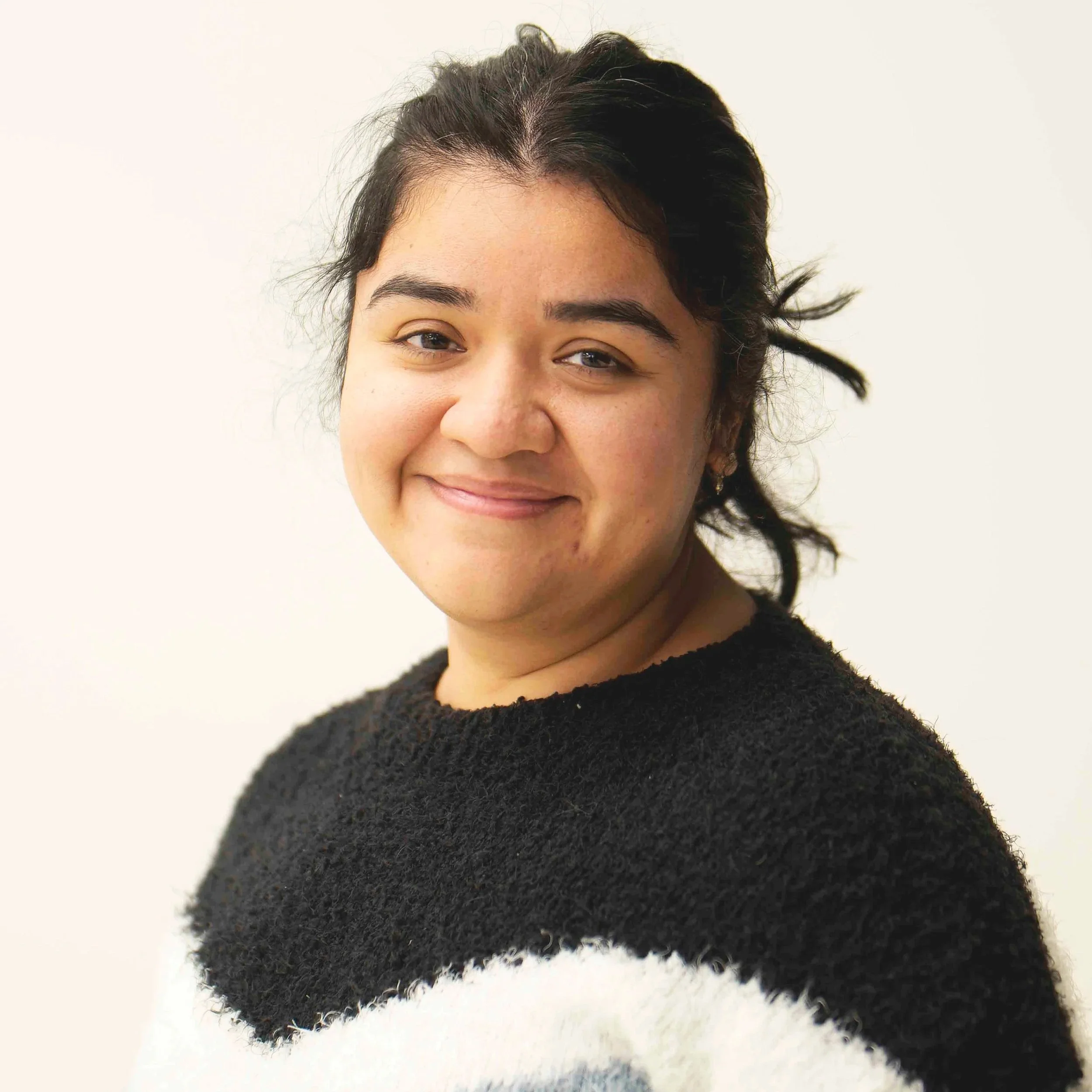 A person with dark hair in a ponytail, wearing a green sweater, smiles slightly while standing against a plain background.