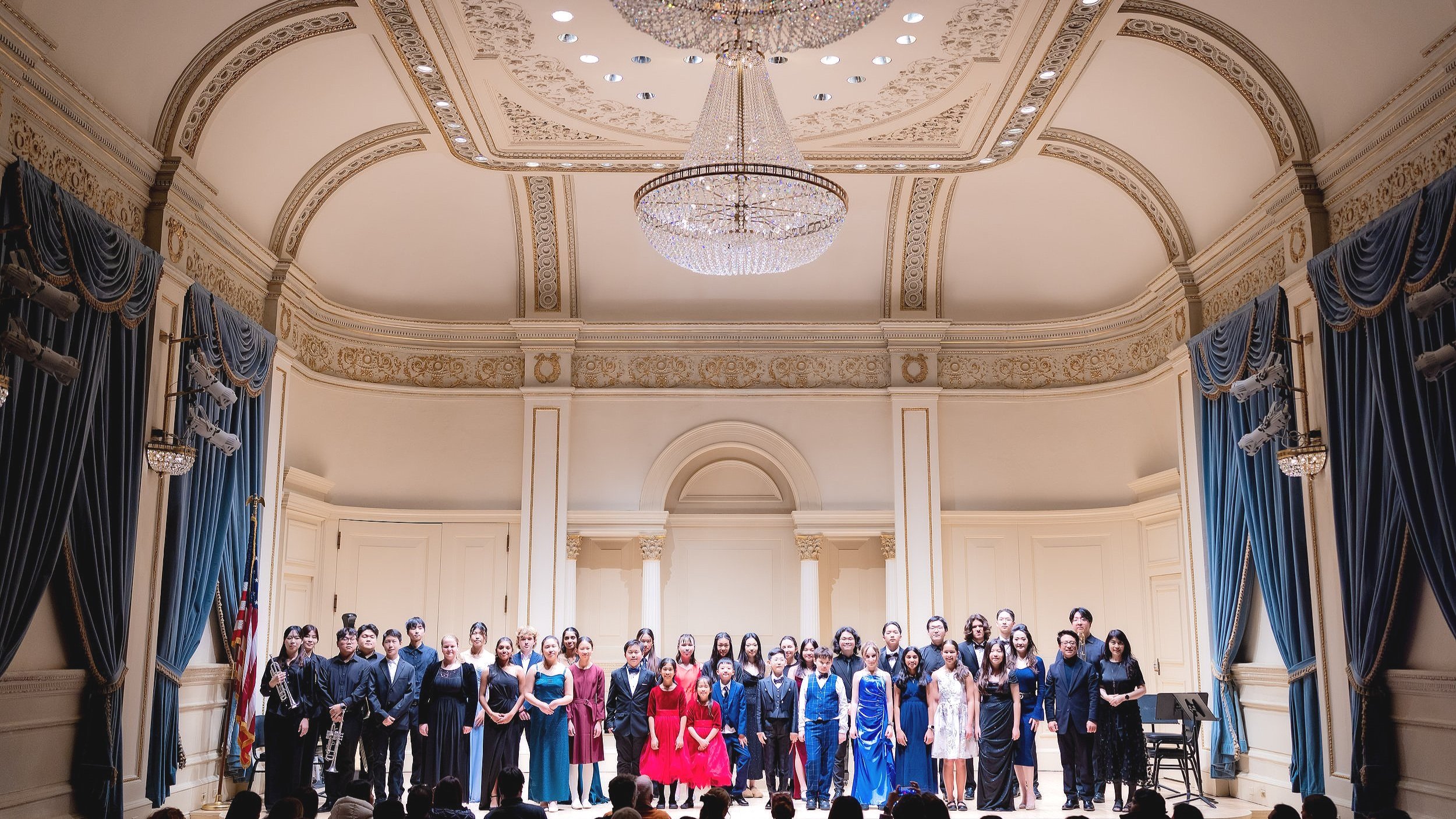 A group of people posing on stage in an ornate concert hall with a chandelier and blue curtains.