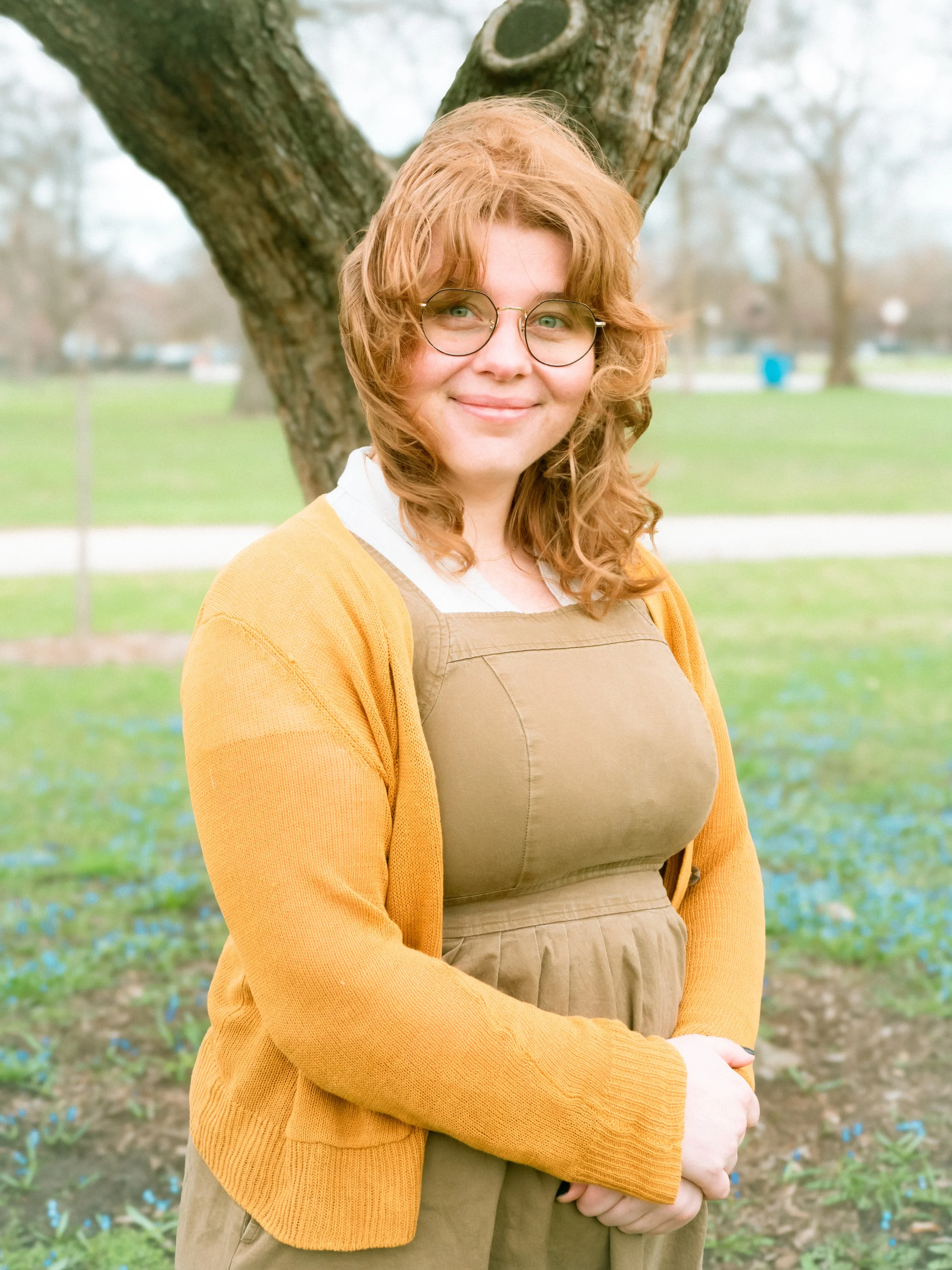 A person with curly red hair and glasses standing outdoors in front of a large tree, smiling, wearing a mustard-colored cardigan and a tan apron.