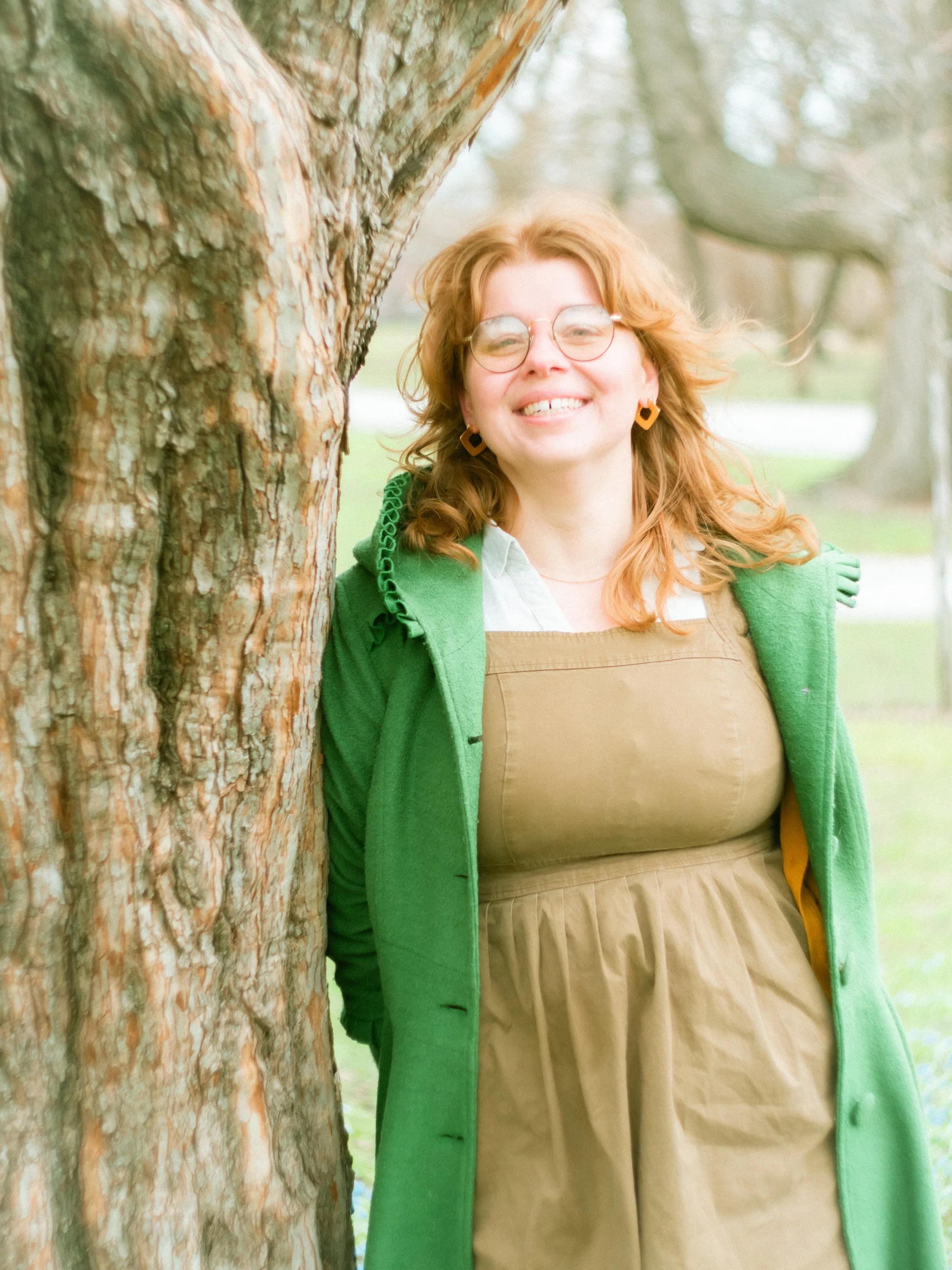 A person with red hair, glasses, and earrings smiling while standing outdoors next to a tree in a park.