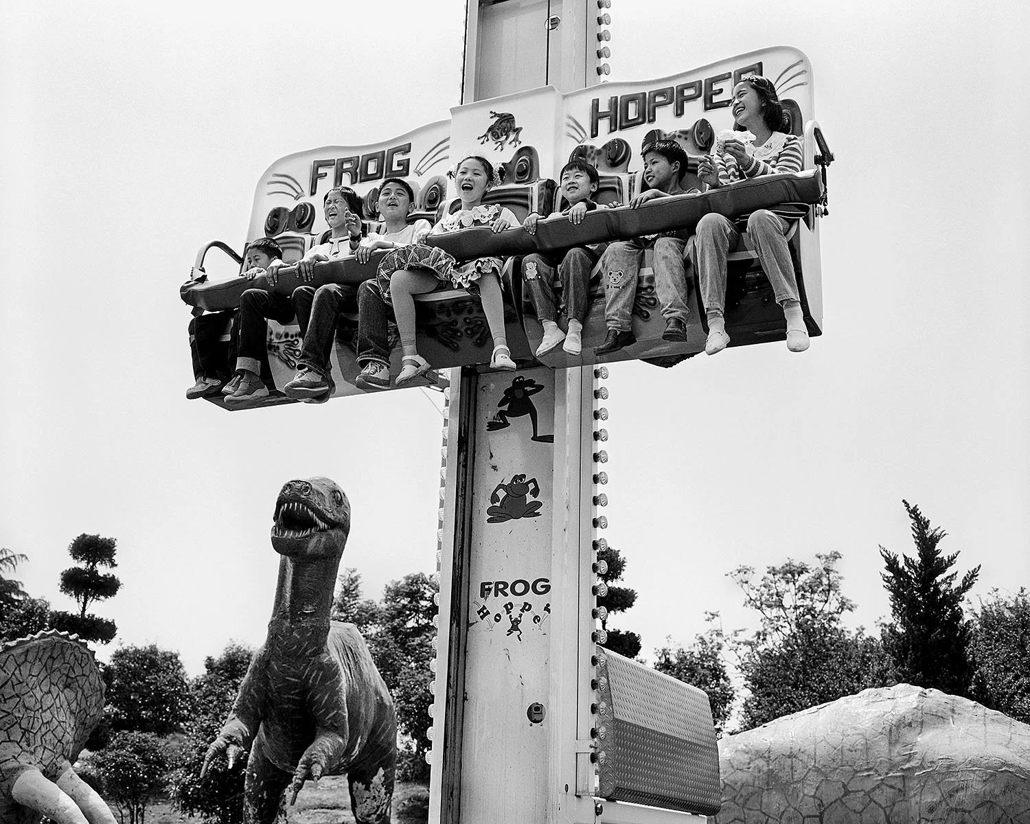 Children laughing on an amusement park ride in Shanghai, 2002, seated above a dinosaur figure that appears to wanting to eat them.