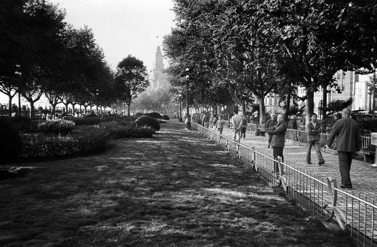 People standing and walking along a tree-lined park edge in Shanghai, 1993, with the Park Hotel visible in the background.