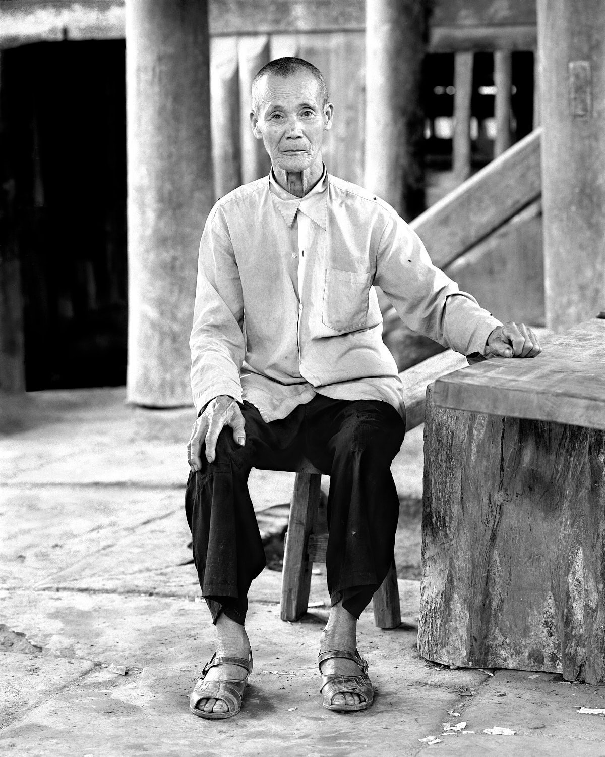 1987, Chengyang, China: an elder wearing sandals sits on a low stool beside a wooden table, with timber columns and steps behind him.