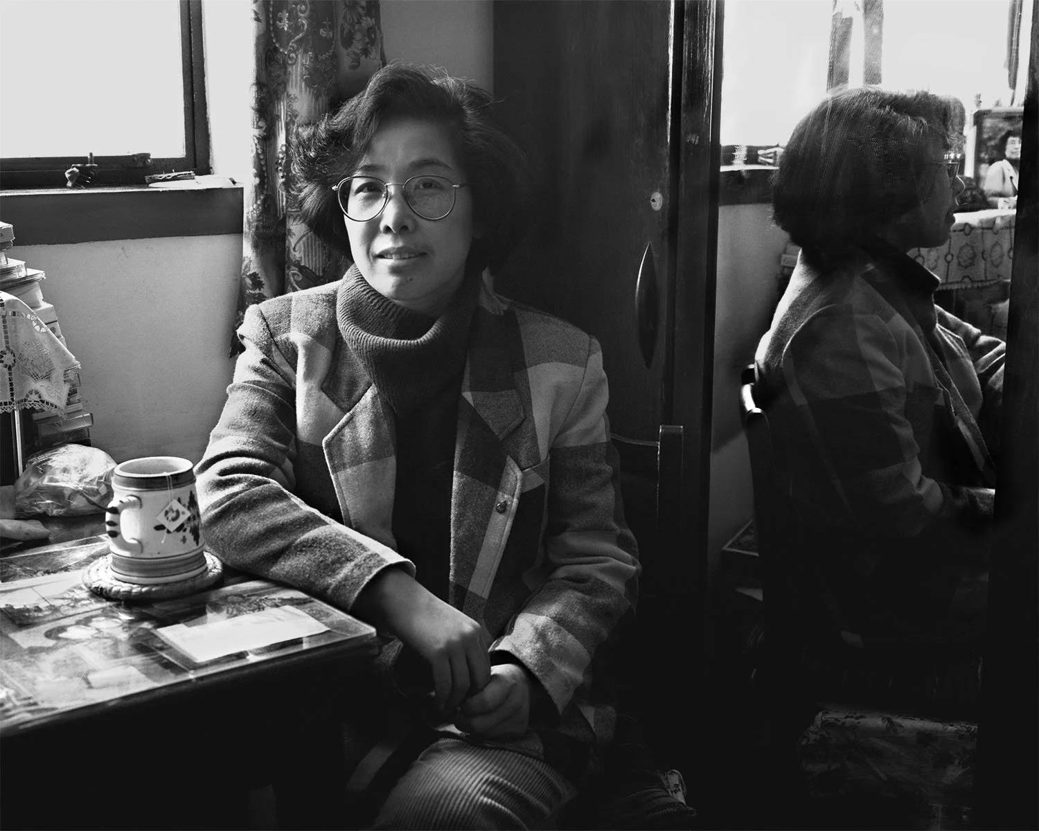 A monochrome portrait of Lin Er seated indoors close to a window in Shanghai, 1988, softly illuminated, with relaxed hands and a contemplative expression.