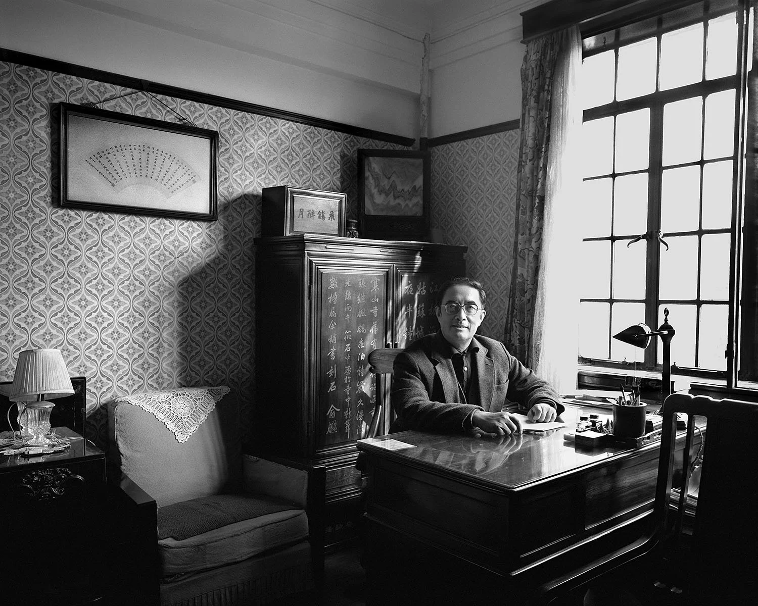 2000, Shanghai: Liu Dalin sits at a polished wooden desk beside a tall window, surrounded by patterned wallpaper, framed artwork, a cabinet, and an armchair.