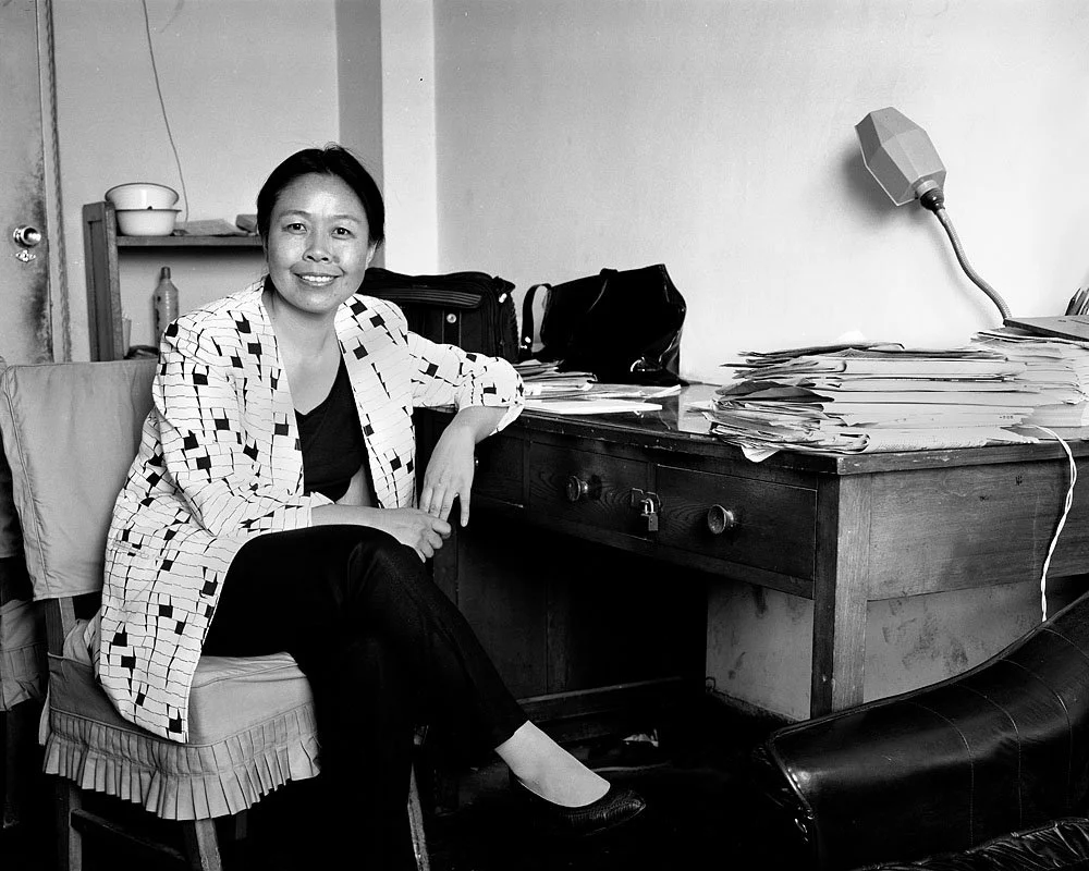 2000, Beijing, China: Dai Qing sits beside a curved wooden desk covered with stacks of papers, with a desk lamp above and one arm resting on the desktop.