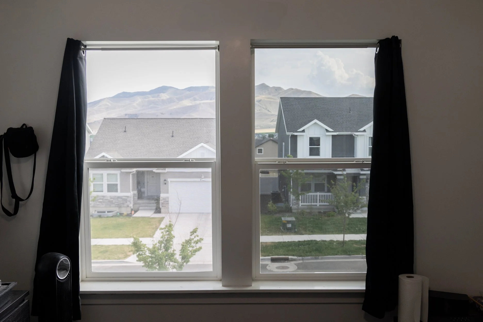 View through two windows showing neighboring houses and mountains in the background, with black curtains and a paper towel roll nearby.