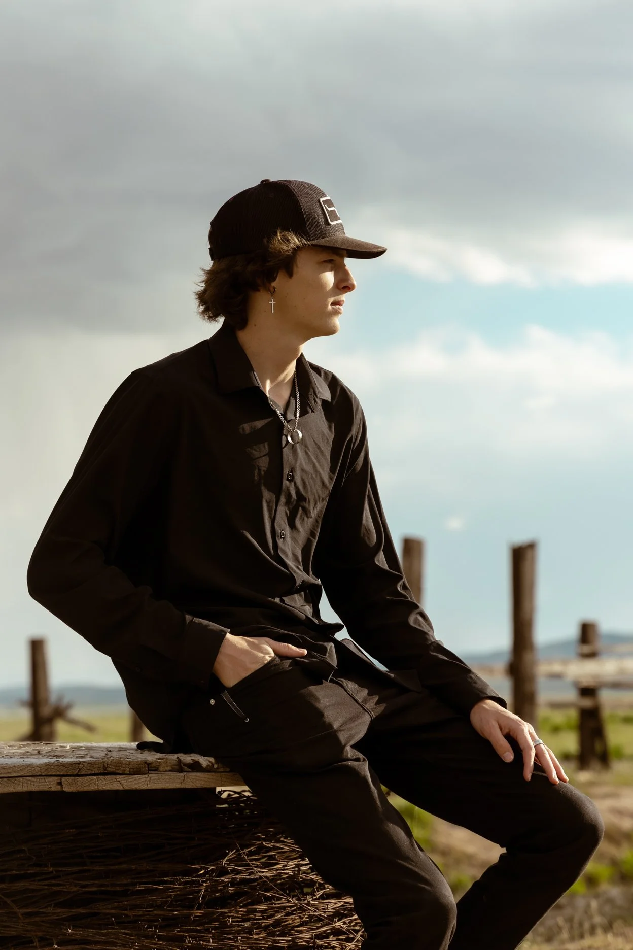 A young man sitting outdoors on a wooden bench, wearing a black shirt, black pants, a black cap, and silver jewelry, with a cloudy sky in the background.