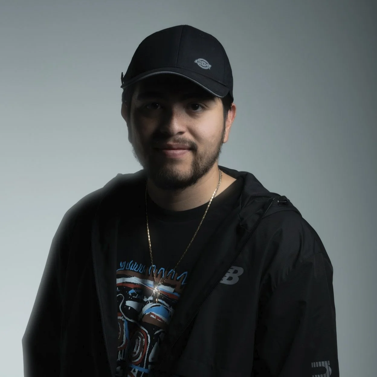 Portrait of a young man with a beard, wearing a black cap, black jacket, and graphic T-shirt, against a plain background.