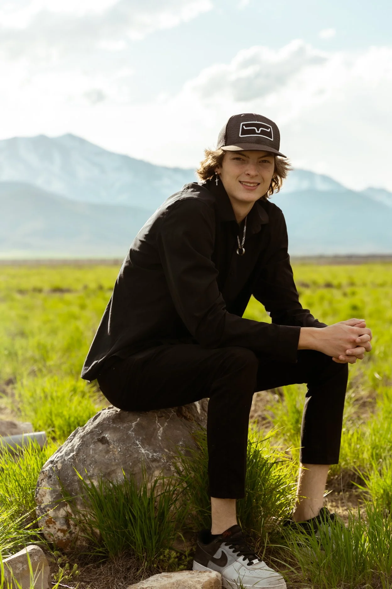 A young person with medium-length hair, wearing a black cap, black shirt and pants, sitting on a rock in a green field with mountains in the background.