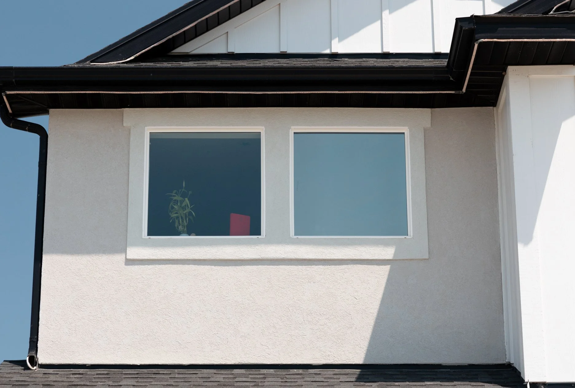 Close-up of a house's exterior with two side-by-side windows, a beige stucco wall, and a black roof with a gutter