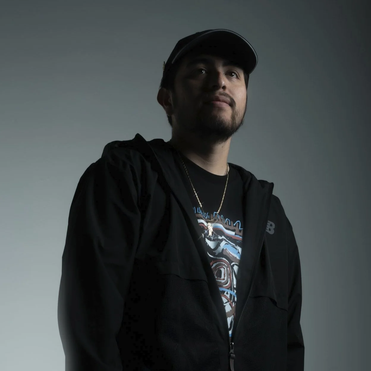 A young man wearing a black jacket, baseball cap, and gold chain against a plain gray background.