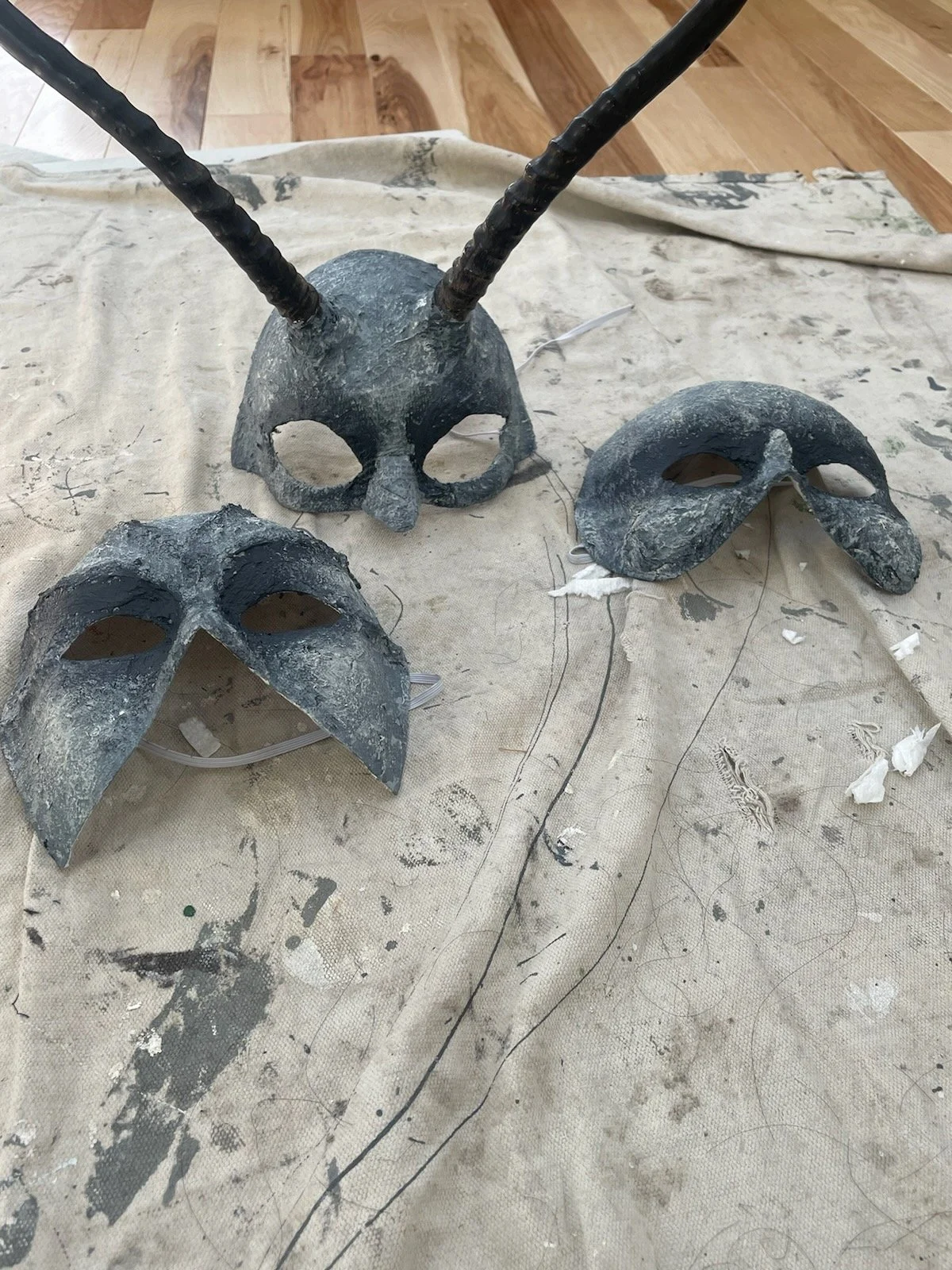 Three witch masks from Macbeth, each unique and painted stony grays and black. One has two antelope antlers attached to the forehead.