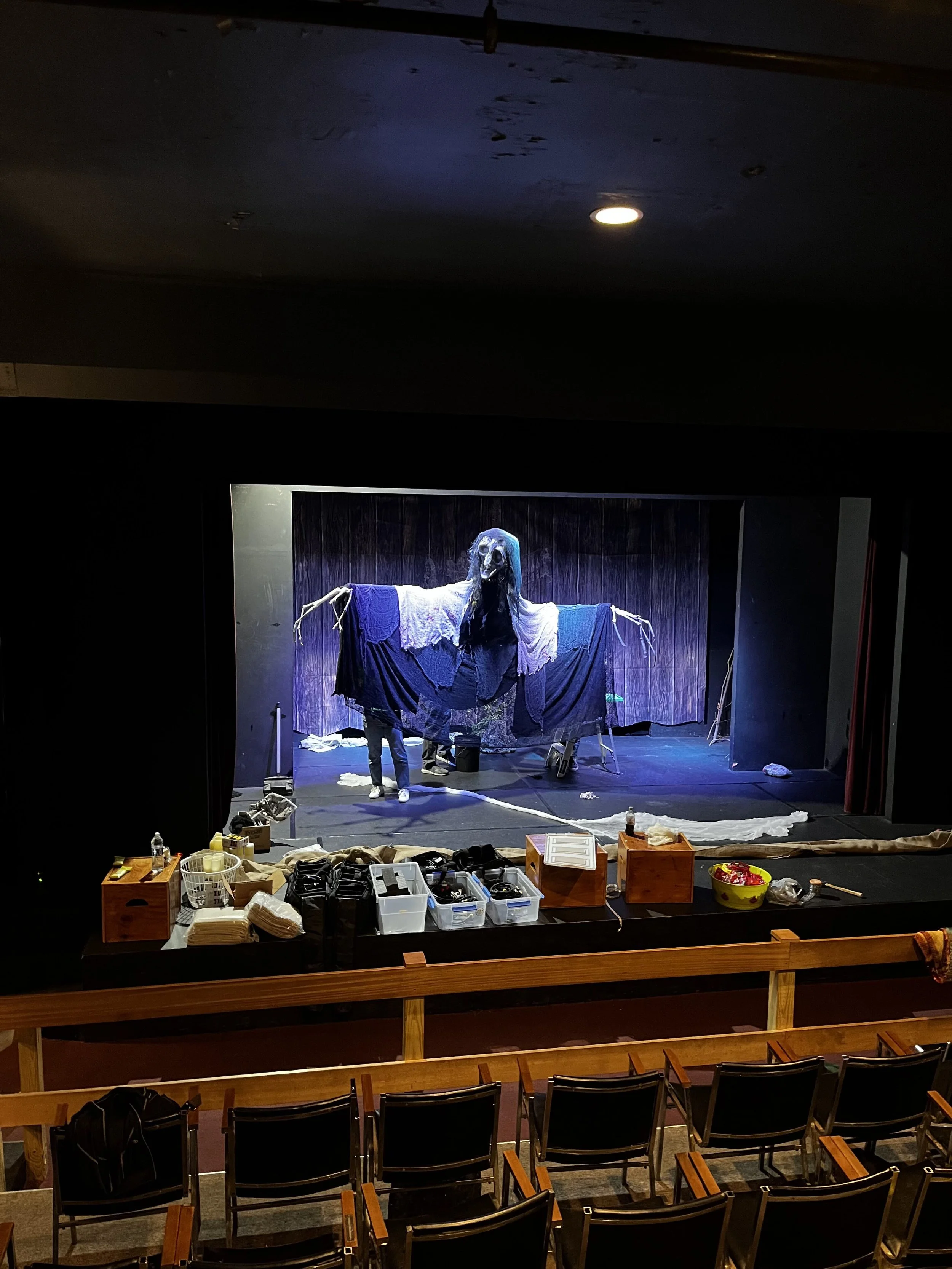 Stage with Hecate puppet being tested during tech week. Large grotesque paper-mache mask, large extended bony fingers, gray and black draping cloth, operated by three people standing behind cloth.