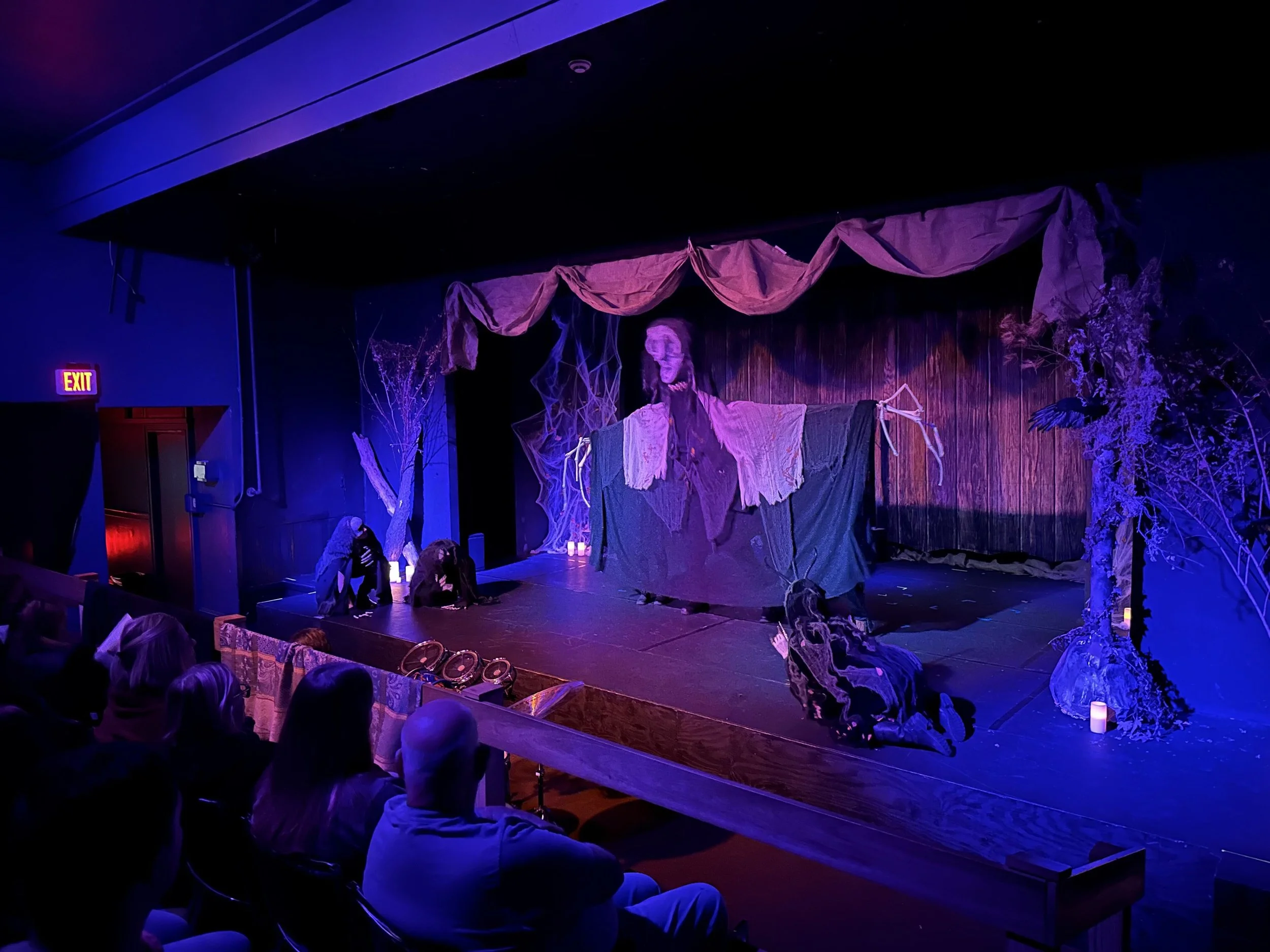 Stage debut of completed Hecate puppet. Blue and red lights washing stage, three witches crouched on edges of stage. Hecate in center, looming over audience seen in bottom left corner.