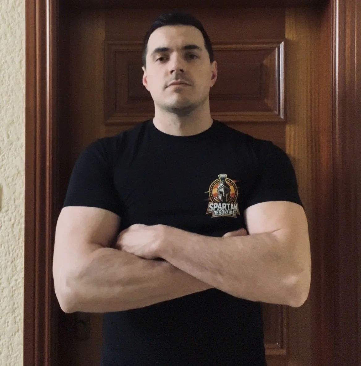 Hombre con brazos cruzados, vestiendo camiseta negra con logotipo del Spartan en el pecho, posando frente a una puerta de madera.