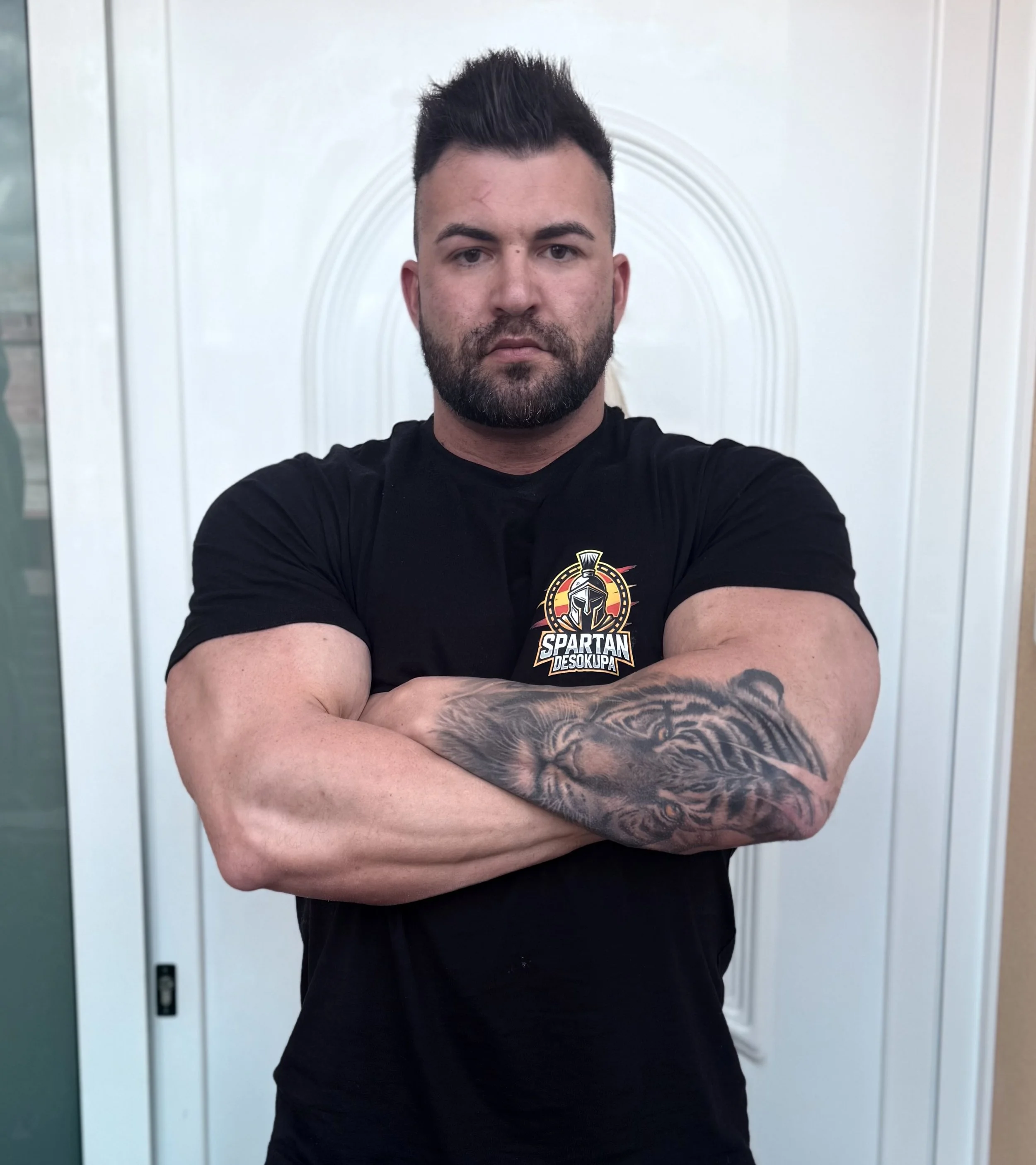 Hombre con barba y peinado hacia arriba, cruzado de brazos, con tatuaje de un tigre en el brazo derecho, vestido con camiseta negra con logo de Spartán Desokupa, frente a puerta blanca con arco decorativo.