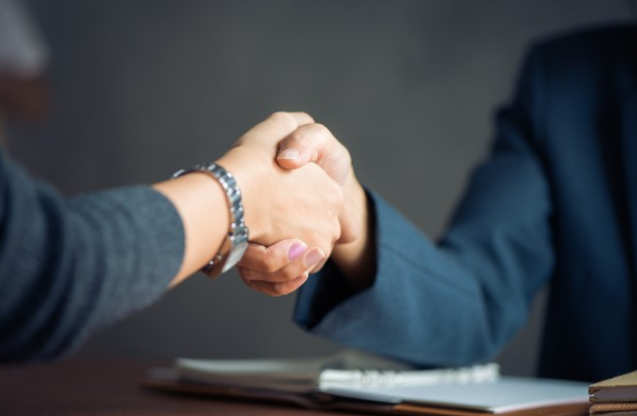 Dos personas dándose la mano en un apretón formal, con un escritorio y documentos en el fondo.