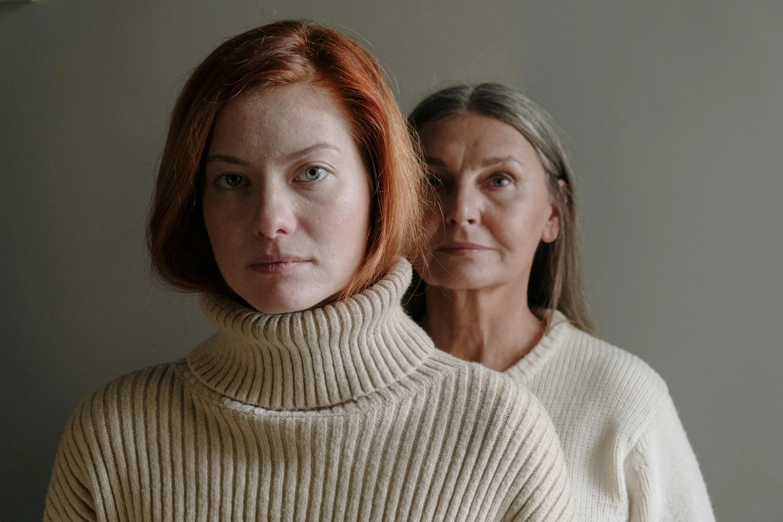 Two women standing close together, both wearing cream sweaters, with one having red hair and the other with gray hair.