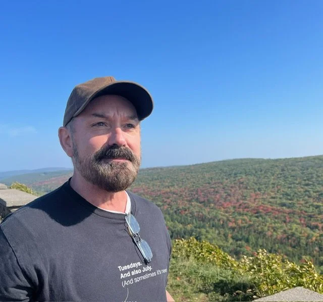 A man with a beard wearing a baseball cap and sunglasses on his shirt stands outdoors against a backdrop of rolling hills and a clear blue sky.