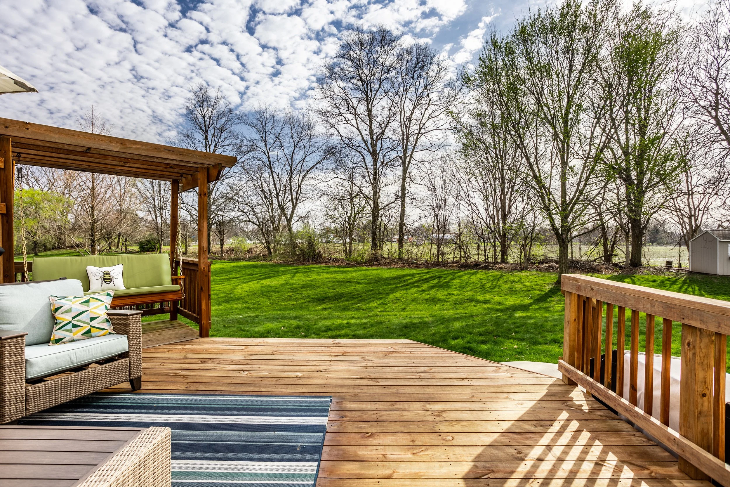 Wooden deck with outdoor seating, blue striped rug, and gazebo; grassy yard and trees in background; clear sky with clouds.