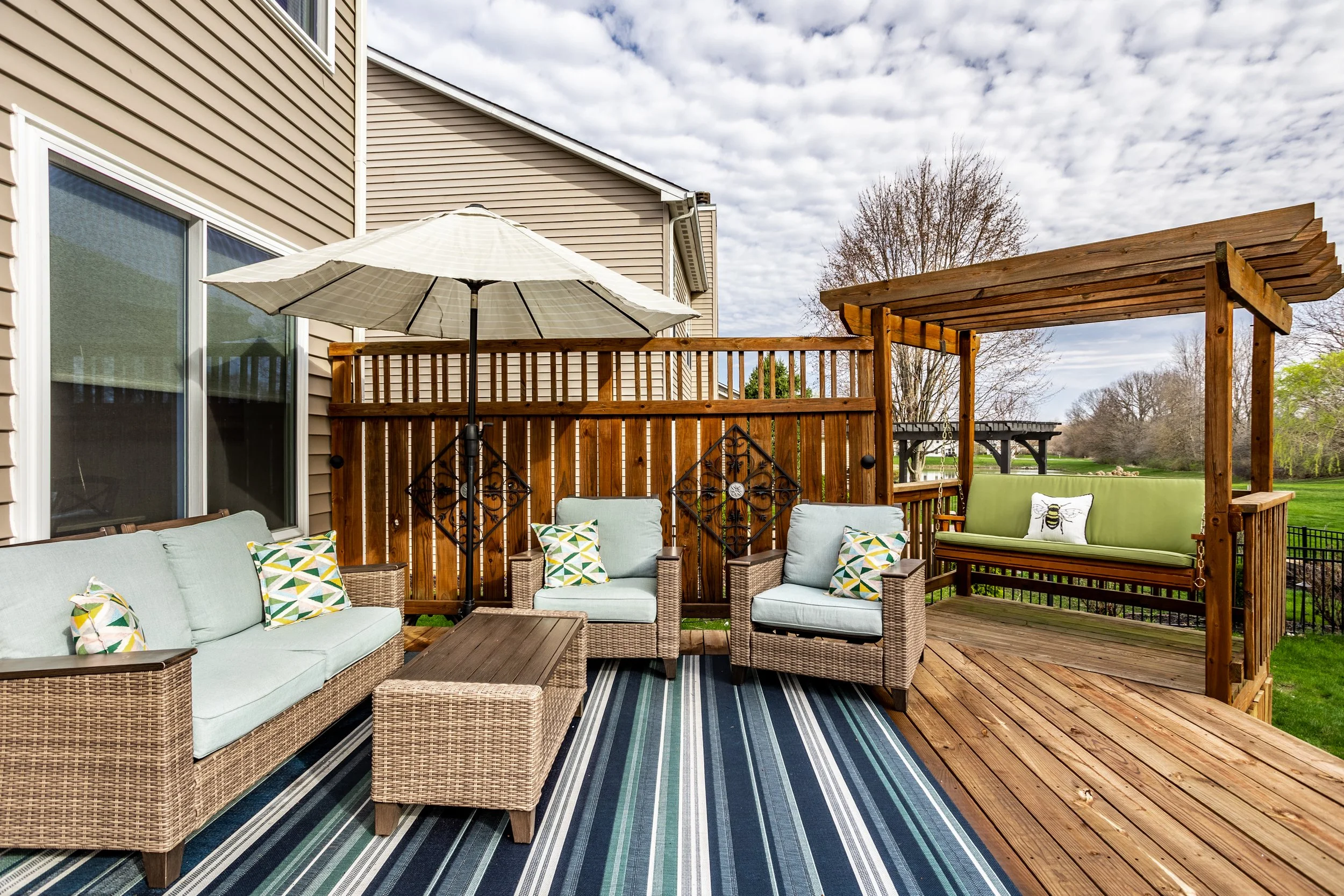 Outdoor patio with wicker furniture, striped rug, umbrella, and wooden pergola with swing.