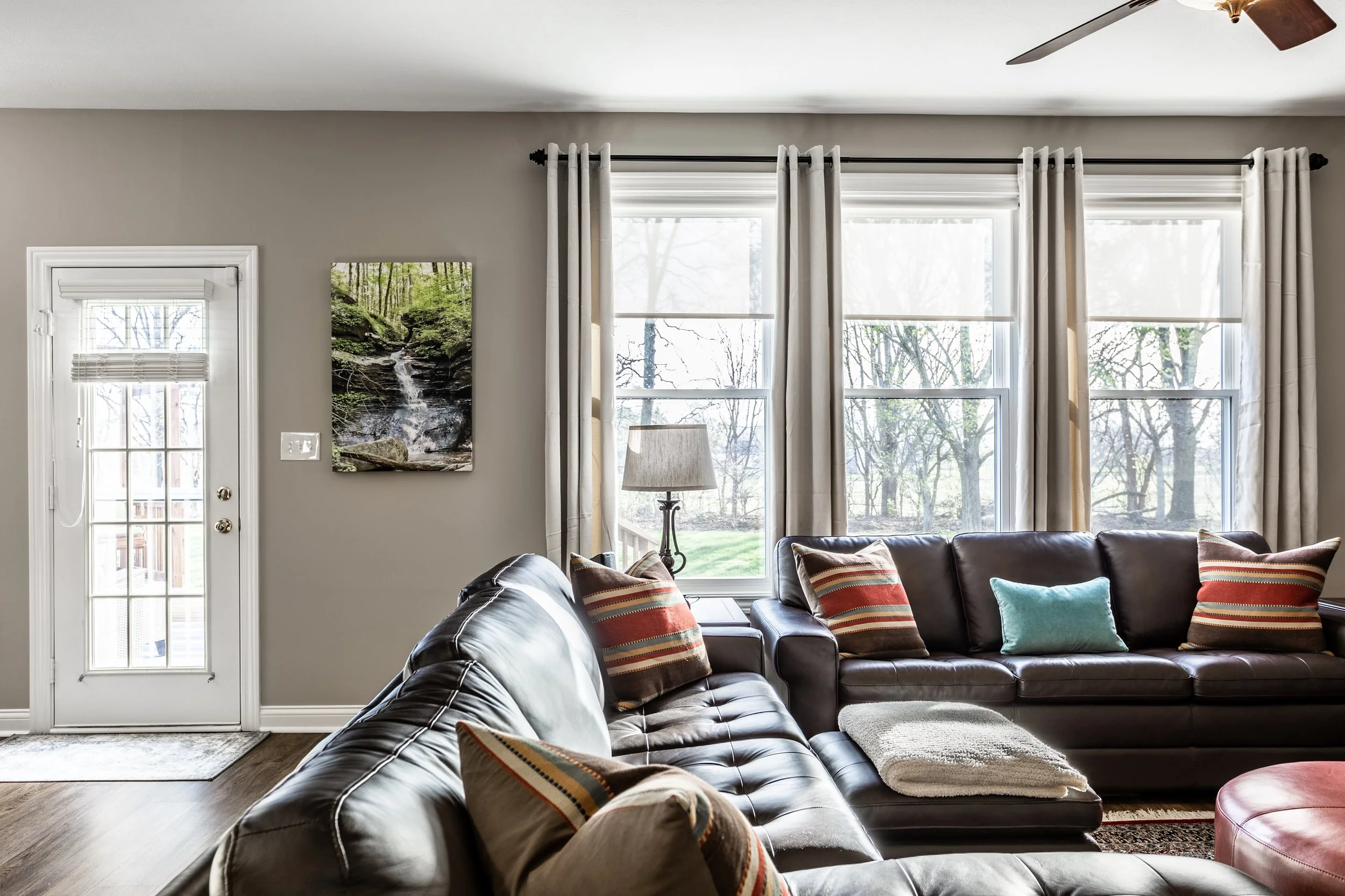 Cozy living room with leather sofas, colorful pillows, and a blanket by large windows. A landscape painting hangs on the wall, and a ceiling fan is visible above.