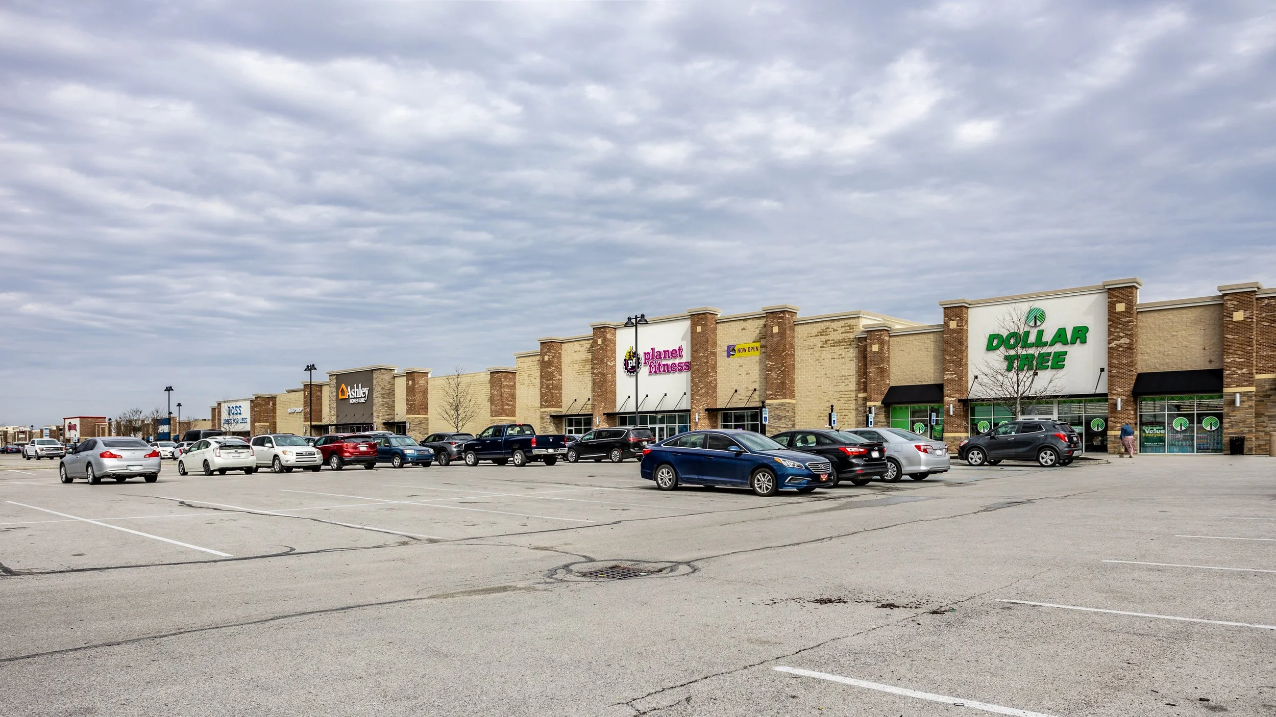Shopping center with Dollar Tree, Planet Fitness, and Ashley HomeStore under cloudy sky, parking lot with several cars.