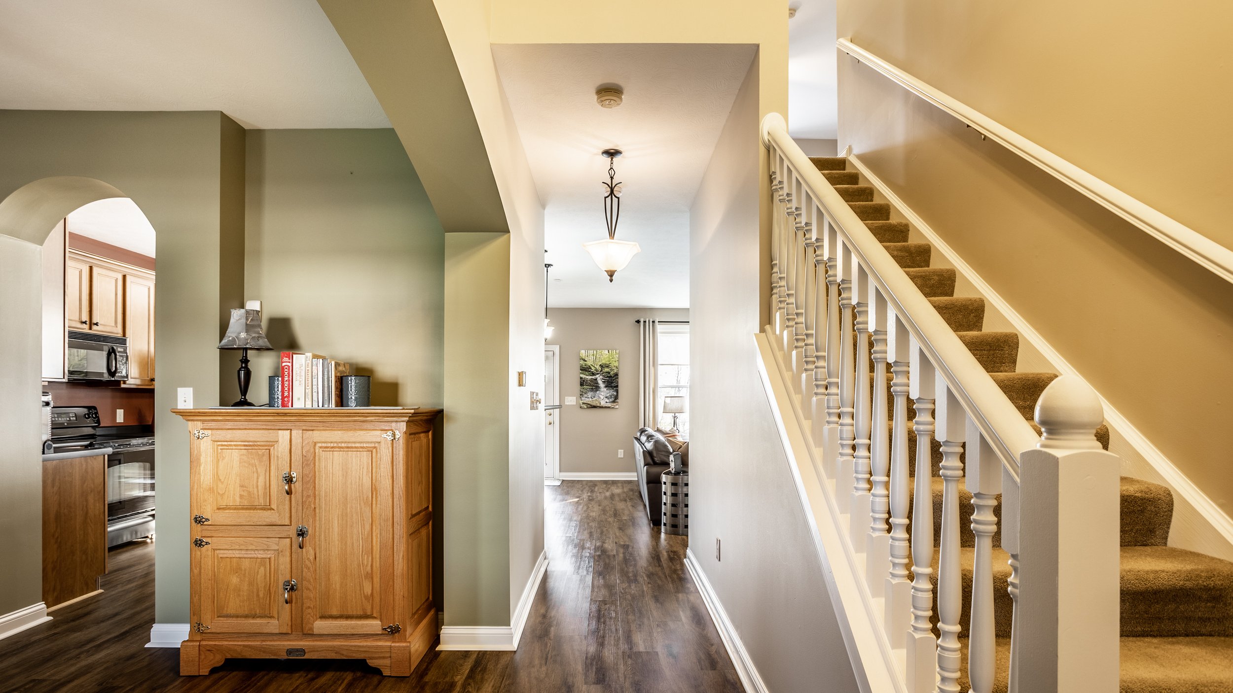 Interior of a home with a wood-floored hallway leading to a living area, a wooden cabinet on the left, and a carpeted staircase on the right.