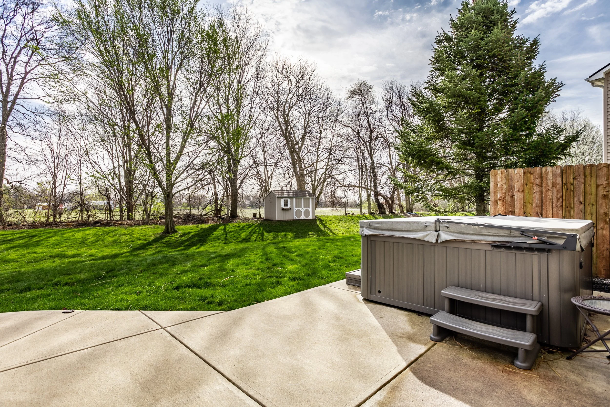 Backyard featuring a hot tub on a concrete patio, lush green grass, mature trees, a wooden fence, and a small storage shed.