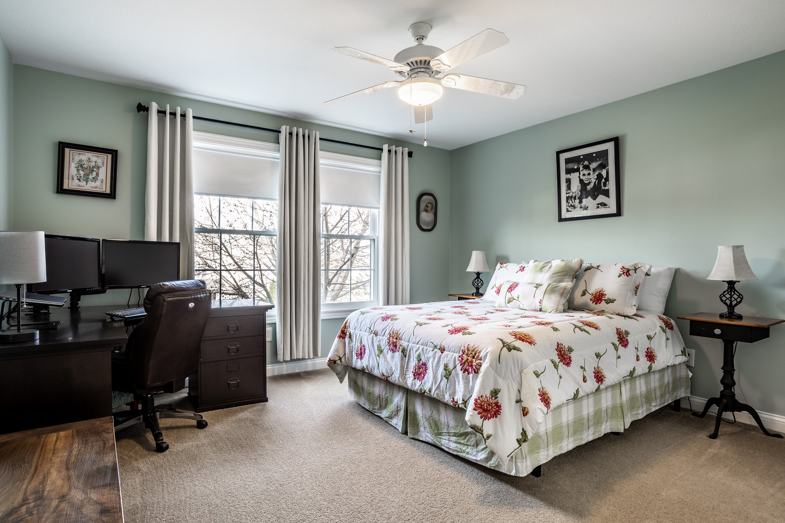Bedroom with green walls, a bed with floral bedding, nightstands with lamps, a desk with dual monitors, and a ceiling fan.