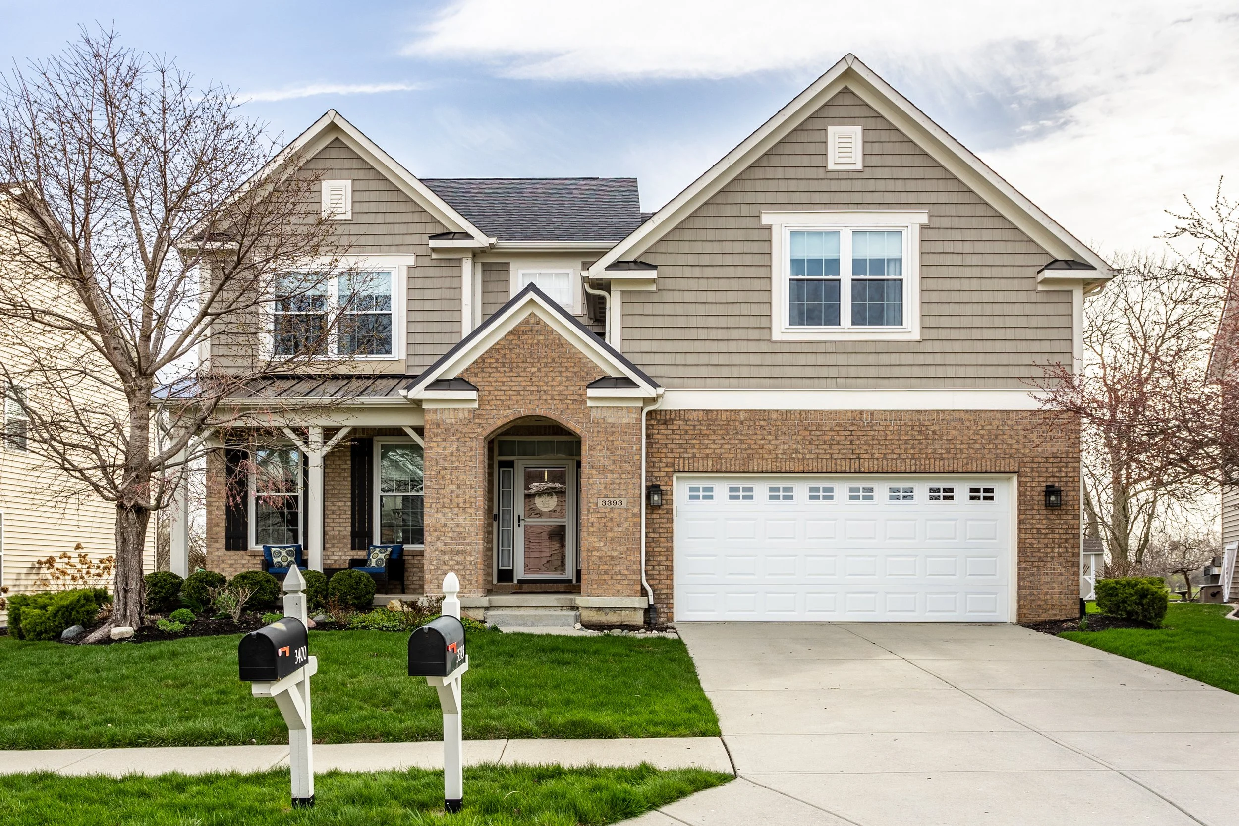 Suburban house with brick and siding exterior, white garage door, two mailboxes, front yard with grass and trees, cloudy sky.