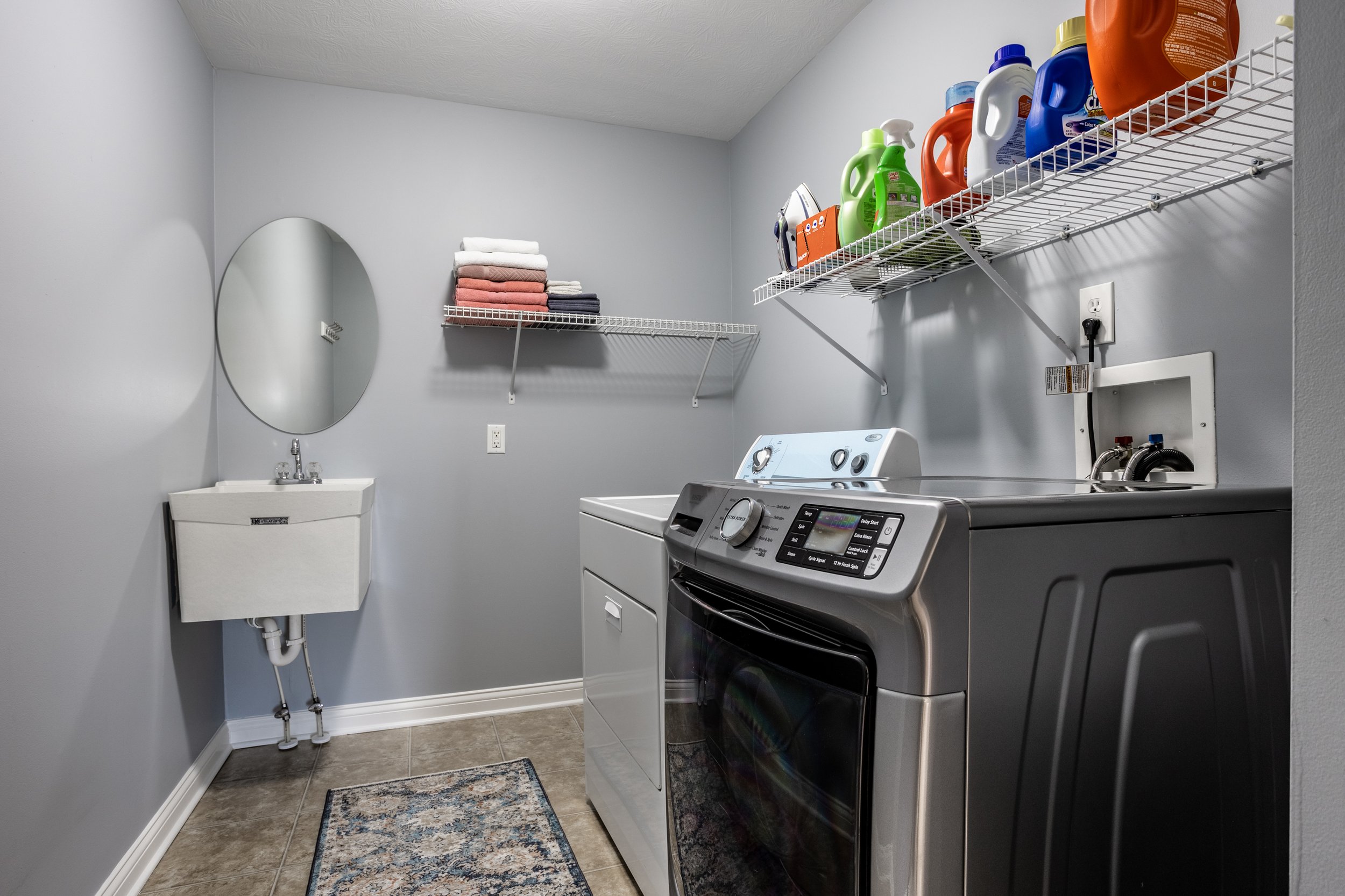 Modern laundry room with washer and dryer, shelves with detergent bottles, folded towels, an iron, and a wall-mounted sink with a round mirror.
