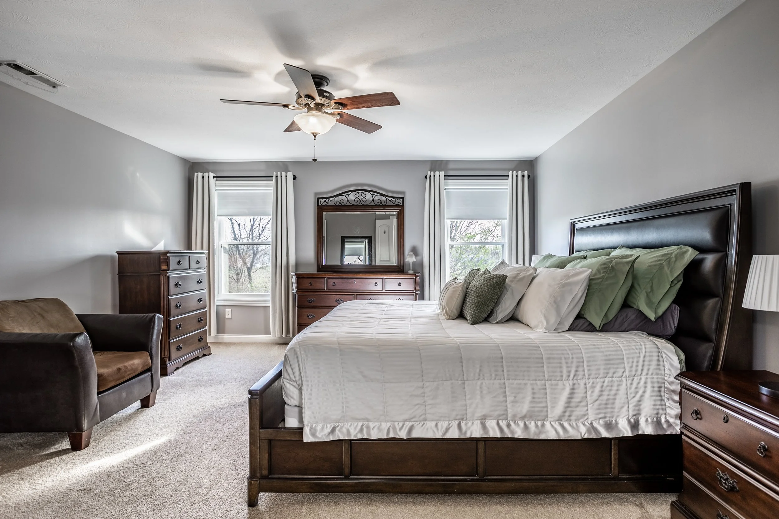 Spacious bedroom with a large bed, multiple pillows, wooden dresser, mirror, armchair, ceiling fan, and windows with white curtains.