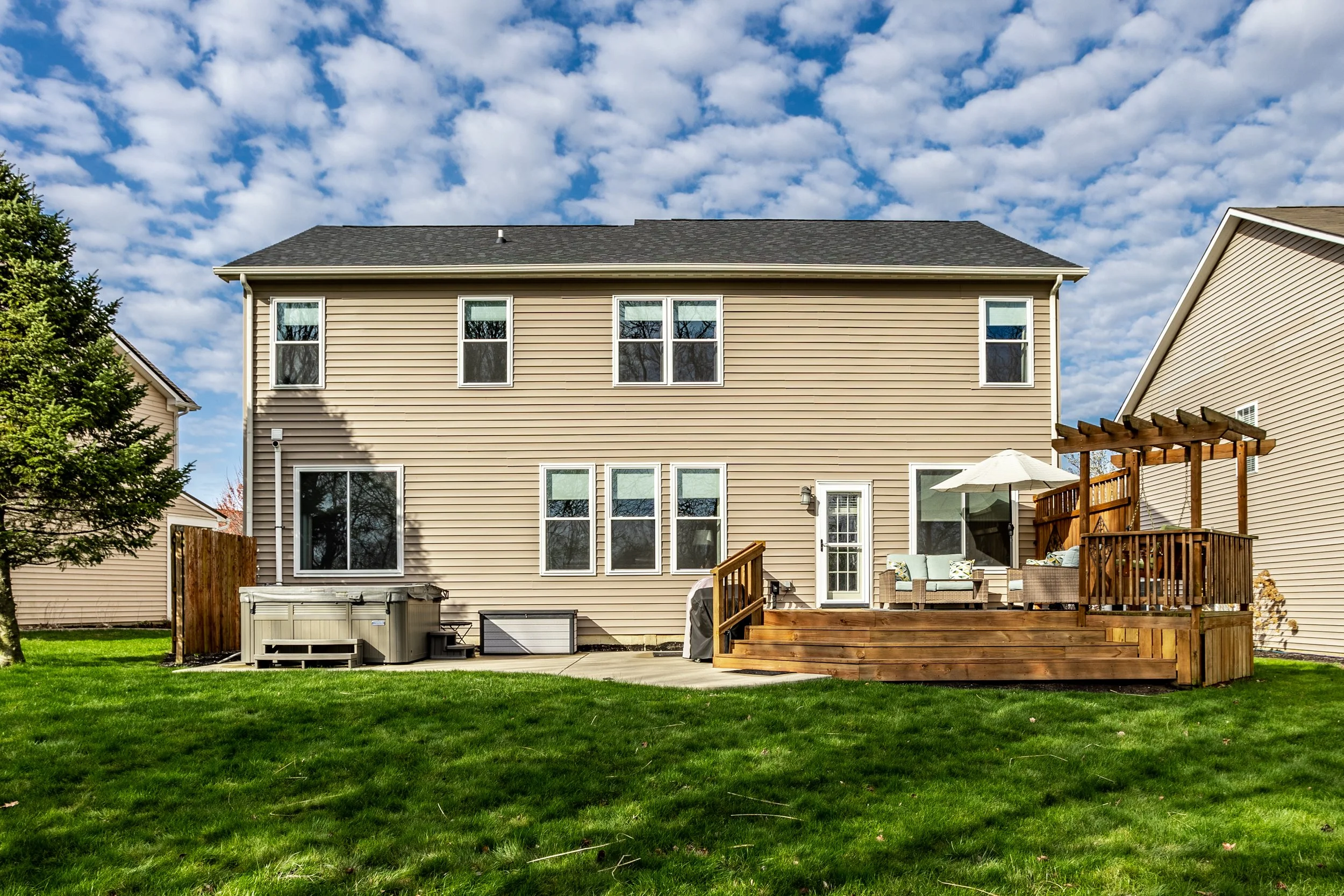 Backyard with two-story beige house, wooden deck, pergola, umbrella, and green grass lawn under a partly cloudy sky.