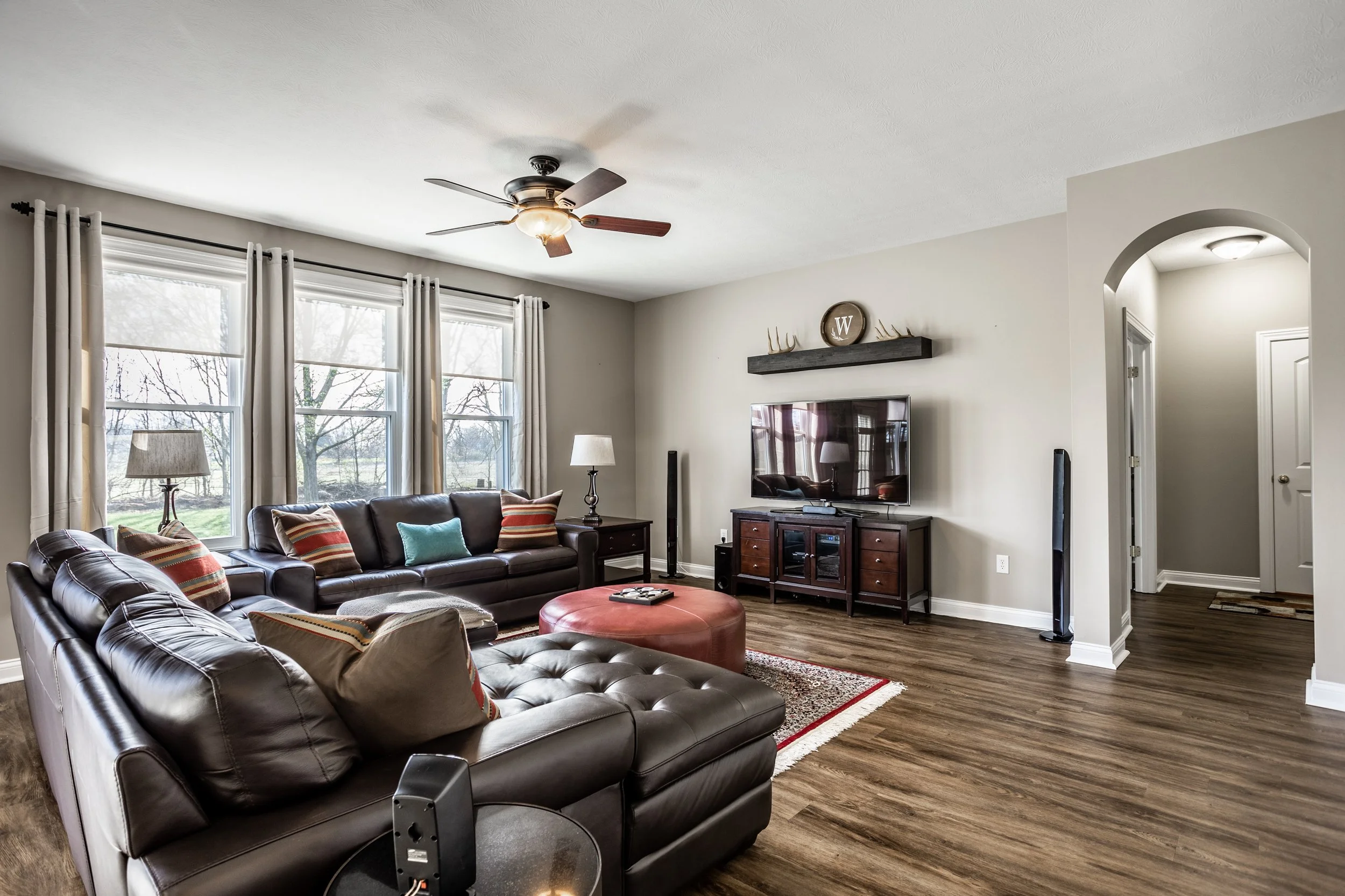 Cozy living room with brown leather sofas, colorful throw pillows, a red ottoman, a ceiling fan, large windows with curtains, a flat-screen TV, wooden TV stand, and a lamp on a side table.