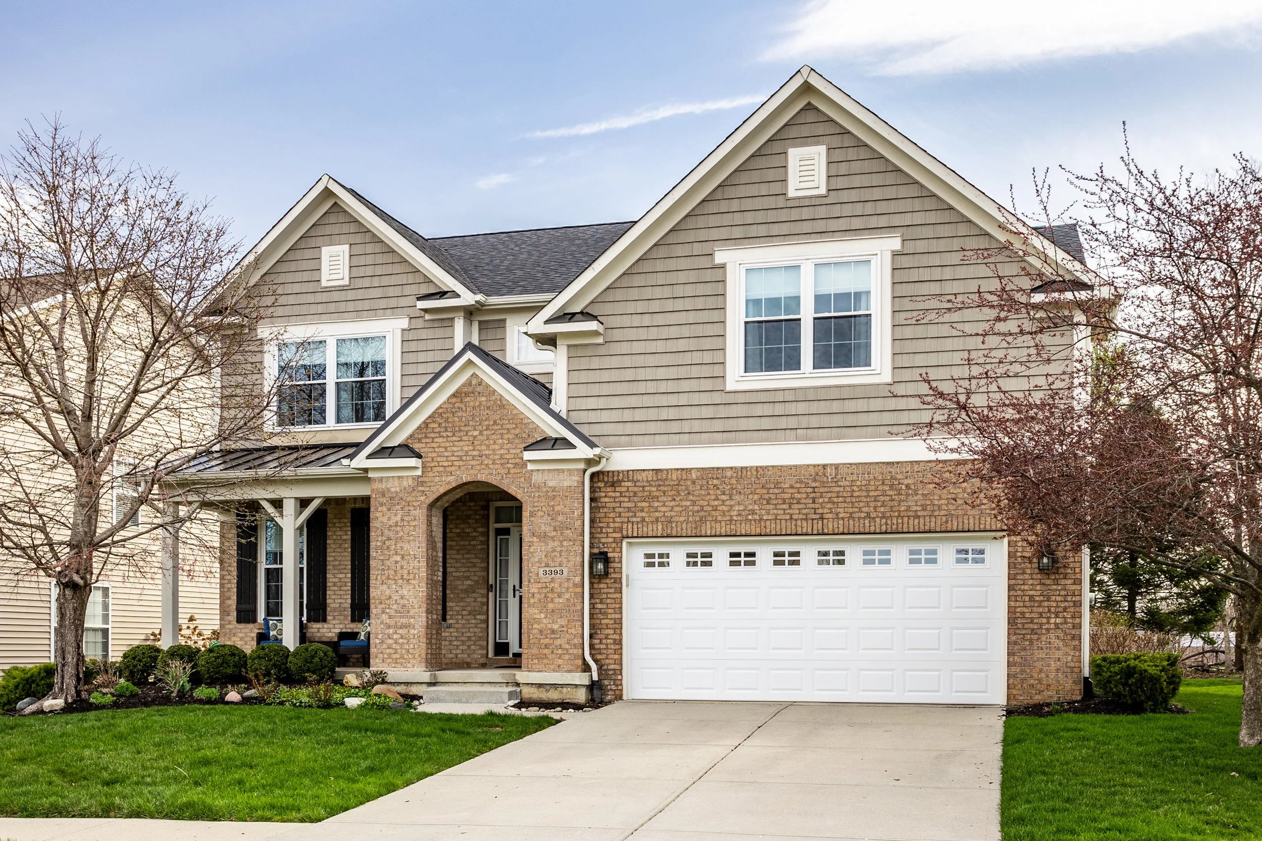 Suburban two-story house with brick facade, white garage door, and landscaped front yard with green grass and bare trees.