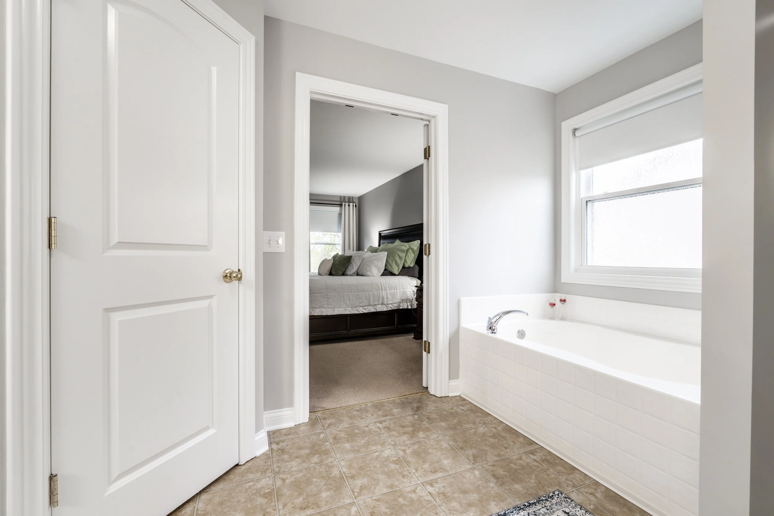 Modern bathroom interior with white bathtub, tile flooring, and doorway leading to a bedroom.