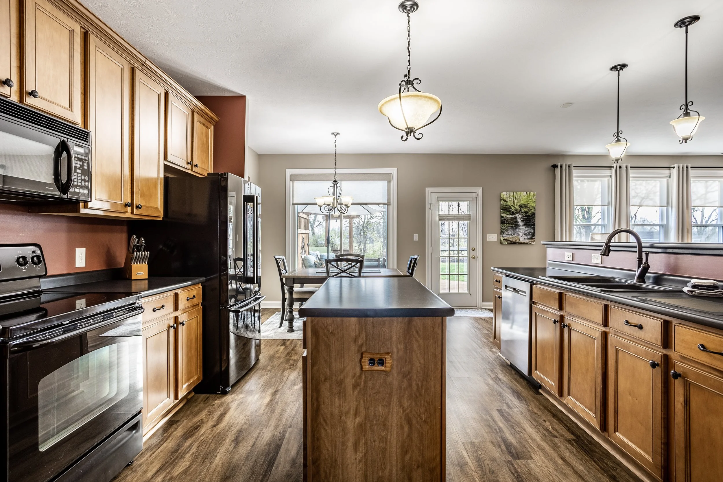 Modern kitchen with wooden cabinets, black appliances, and an island, overlooking a dining area with pendant lighting, wood flooring, and a view of a backyard through large windows and a glass door.