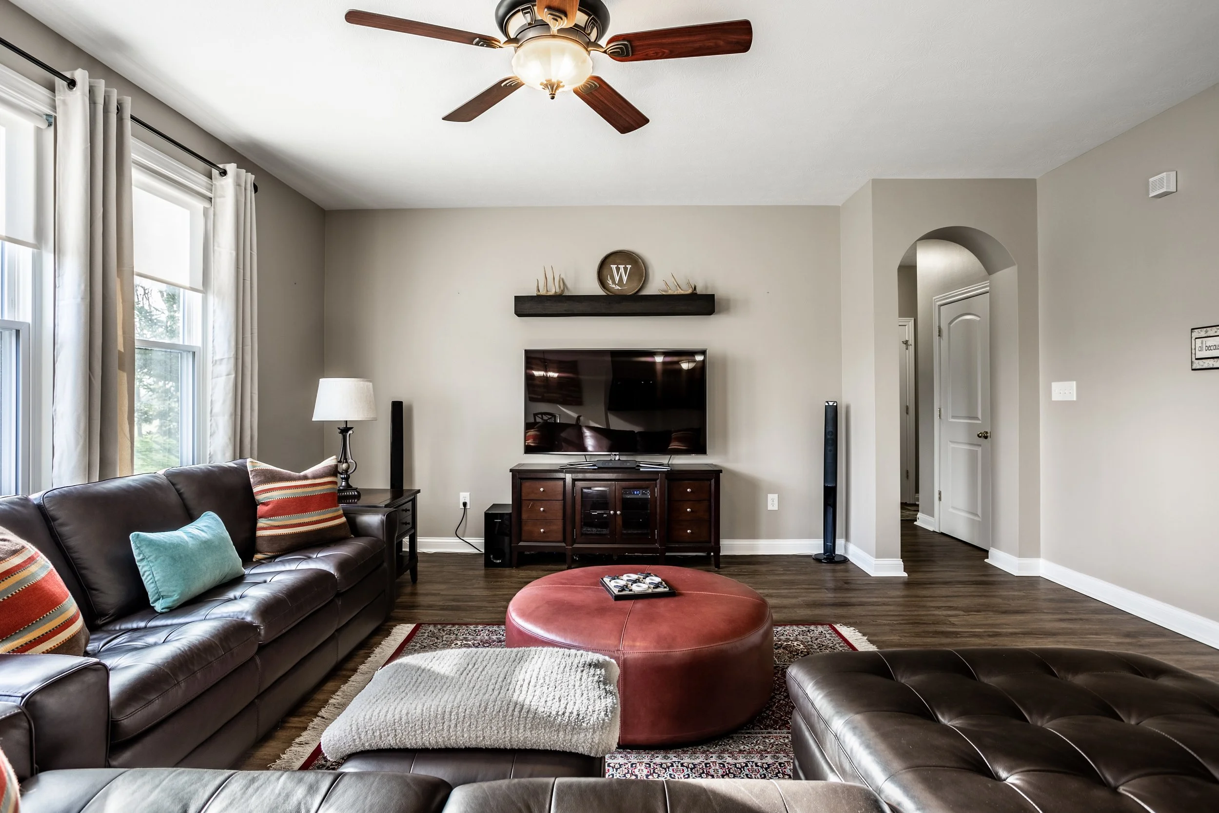 Modern living room with leather sofas, pillows, round ottoman, and TV stand with a flat-screen TV. Ceiling fan, large windows with curtains, wooden floor, and decorative wall shelf. Bright and inviting atmosphere.